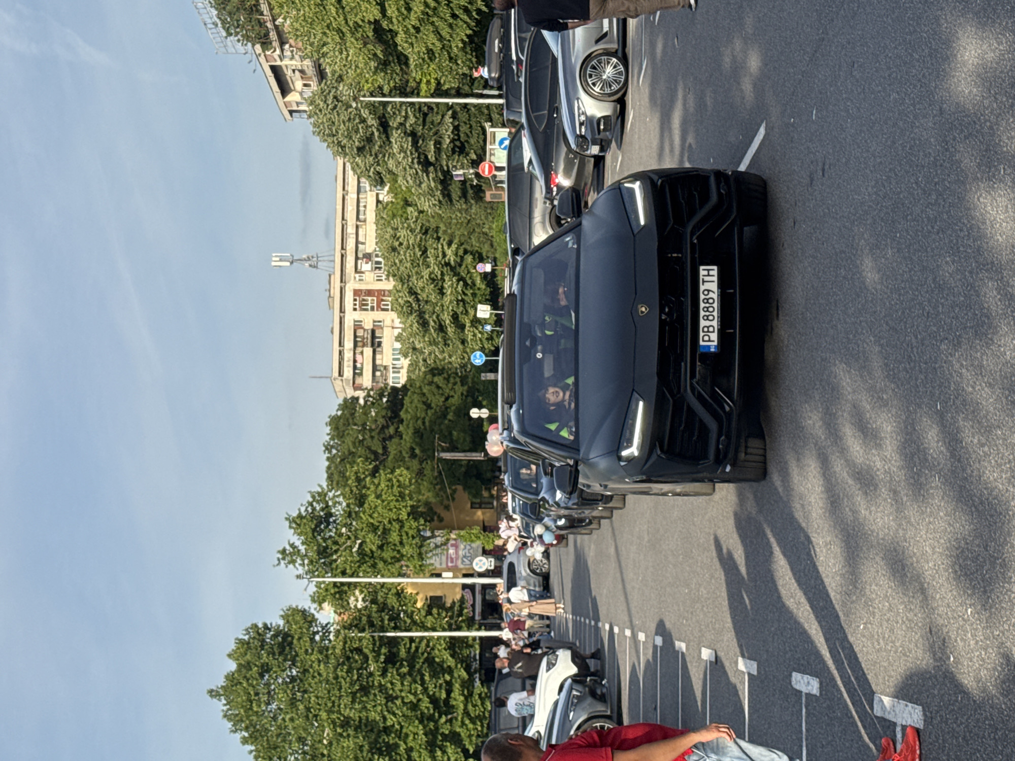 The image depicts a street in Plovdiv, Bulgaria, featuring a line of luxury vehicles parked along the side of the road. The foreground is dominated by a black Lamborghini Urus, license plate "PB 8889 TH." Other high-end vehicles are visible in the background, including what appears to be a silver sports car. A group of people is gathered on the sidewalk, suggesting a possible event or gathering. The environment includes trees and buildings, typical of an urban setting in Plovdiv.