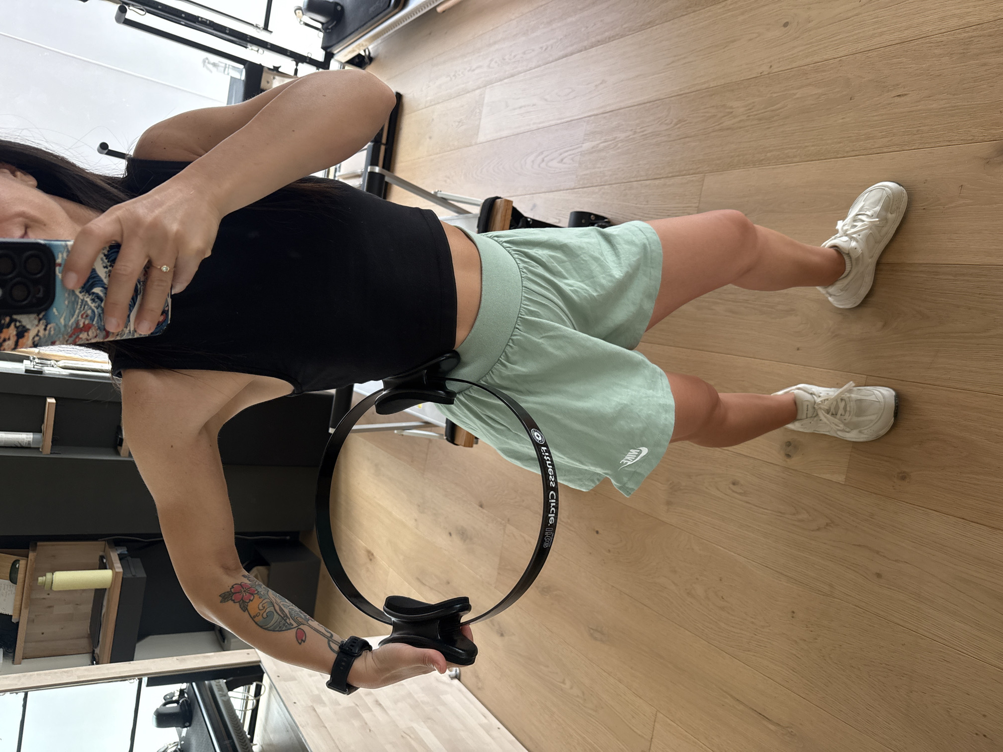 A woman stands in a gym, holding a black pilates ring. She is wearing a black crop top, light green shorts, and white sneakers. The gym has wooden floors and mirrors. There is pilates equipment in the background. The woman has a tattoo on her left arm. She is taking a selfie with her phone. The image was taken in Barcelona, Spain.