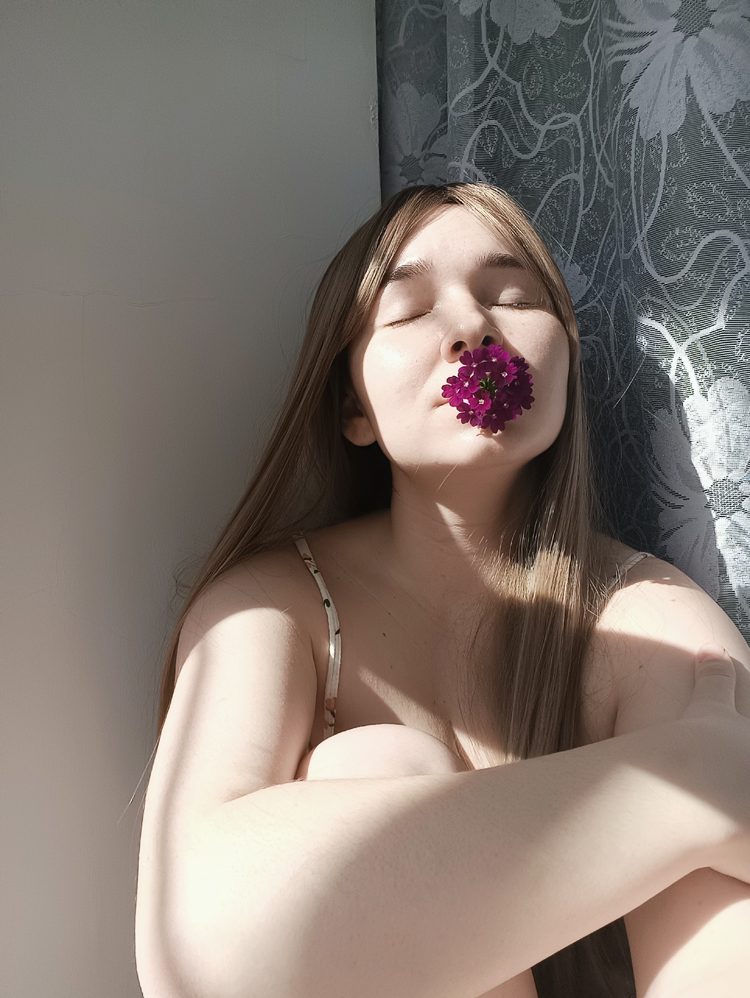 A young, fair-skinned woman with long, light-brown hair is sitting with her eyes closed, holding a purple flower between her nose and mouth. She is leaning against a white wall and a patterned curtain. Her arms are crossed and pulled towards her body. The scene is illuminated by sunlight. The location is not identifiable as Izhevsk, Russia from this image.