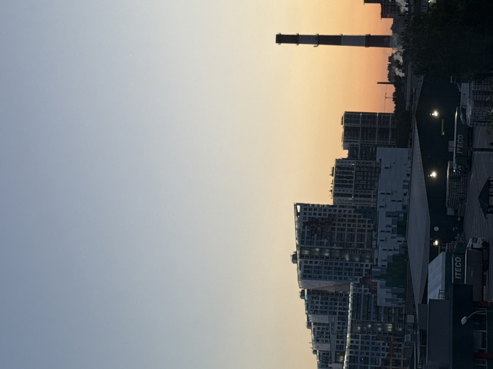 The image, correctly oriented horizontally, presents an urban and industrial landscape captured during either sunrise or sunset. The sky, occupying the left and upper portions, displays a soft gradient, transitioning from a light blue-grey on the far left to a warm orange-yellow on the right horizon. A faint bright spot is visible in the blue-grey area of the sky.
The right side of the scene is filled with a dense cluster of contemporary multi-story buildings, characterized by numerous windows. Some buildings show signs of ongoing construction. In the mid-ground, a prominent industrial chimney, distinct with its red and white stripes, stands against the warm sky, emitting a plume of smoke or steam. Other industrial structures and cranes are visible further in the distance. Dark foliage and trees are interspersed among the buildings and industrial elements.
The lower right foreground features a darker area, likely an industrial yard or road, where several large trucks are visible. Artificial lights from streetlamps or industrial facilities illuminate parts of this area. The word "ITECO" is clearly visible on the sides of at least two of these trucks. The scene conveys the vastness of an urban environment intertwined with industrial activity, under the atmospheric glow of twilight. The location is Saint Petersburg, Russia.