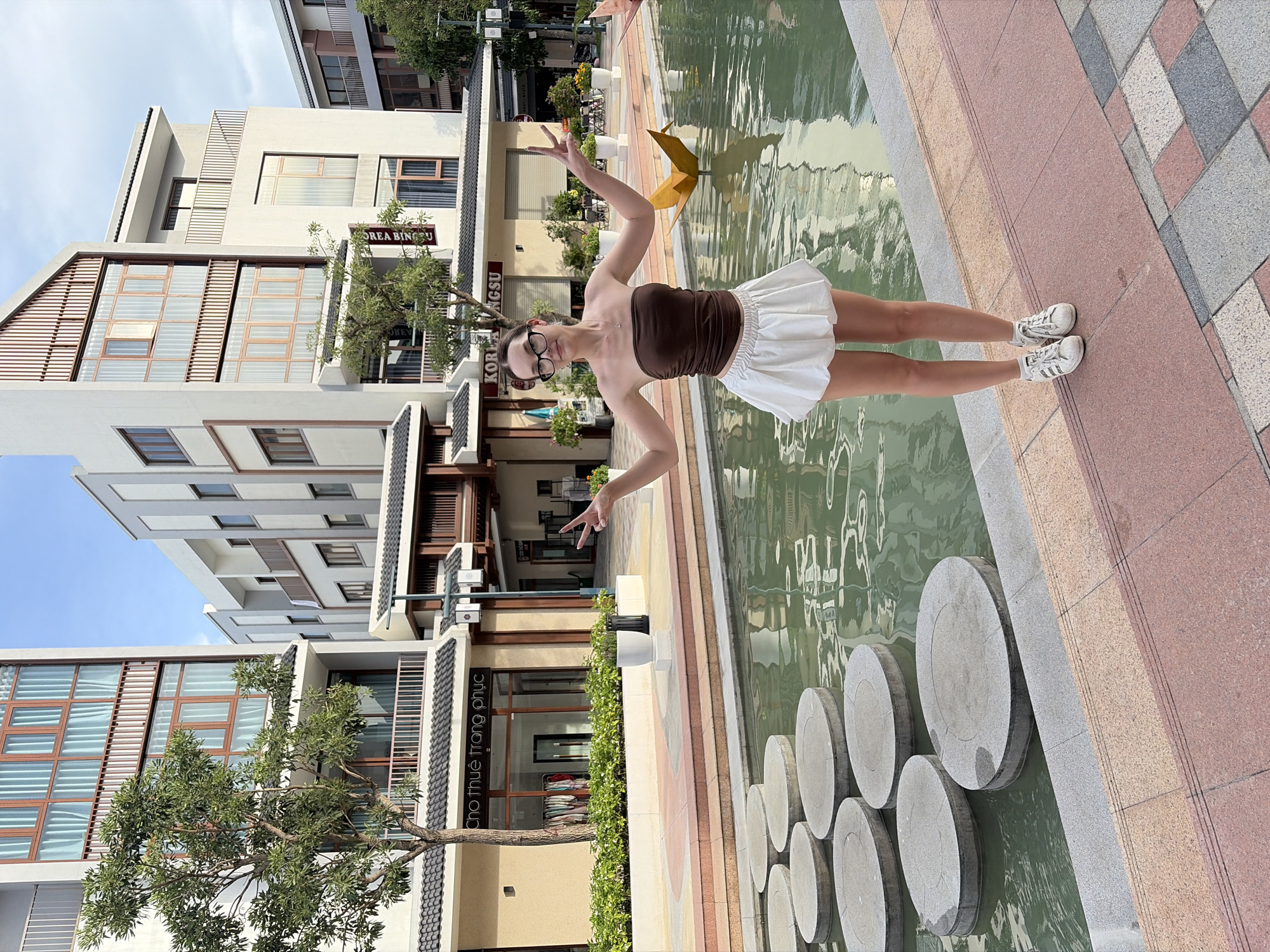 A young Caucasian woman with dark hair pulled back and wearing glasses stands on a paved walkway next to a body of water with circular stepping stones. She is wearing a brown strapless top and a white ruffled skirt, paired with white sneakers. Her arms are raised, and her hands are making peace signs. Behind her, there are modern, multi-story buildings with balconies and large windows. The environment appears to be a commercial or residential area with some greenery and decorative elements. The signage on the buildings suggests a commercial establishment, possibly a shop. The scene is outdoors, during daylight, with a clear sky. The location is identified as Nghĩa Trụ Commune, Vietnam.