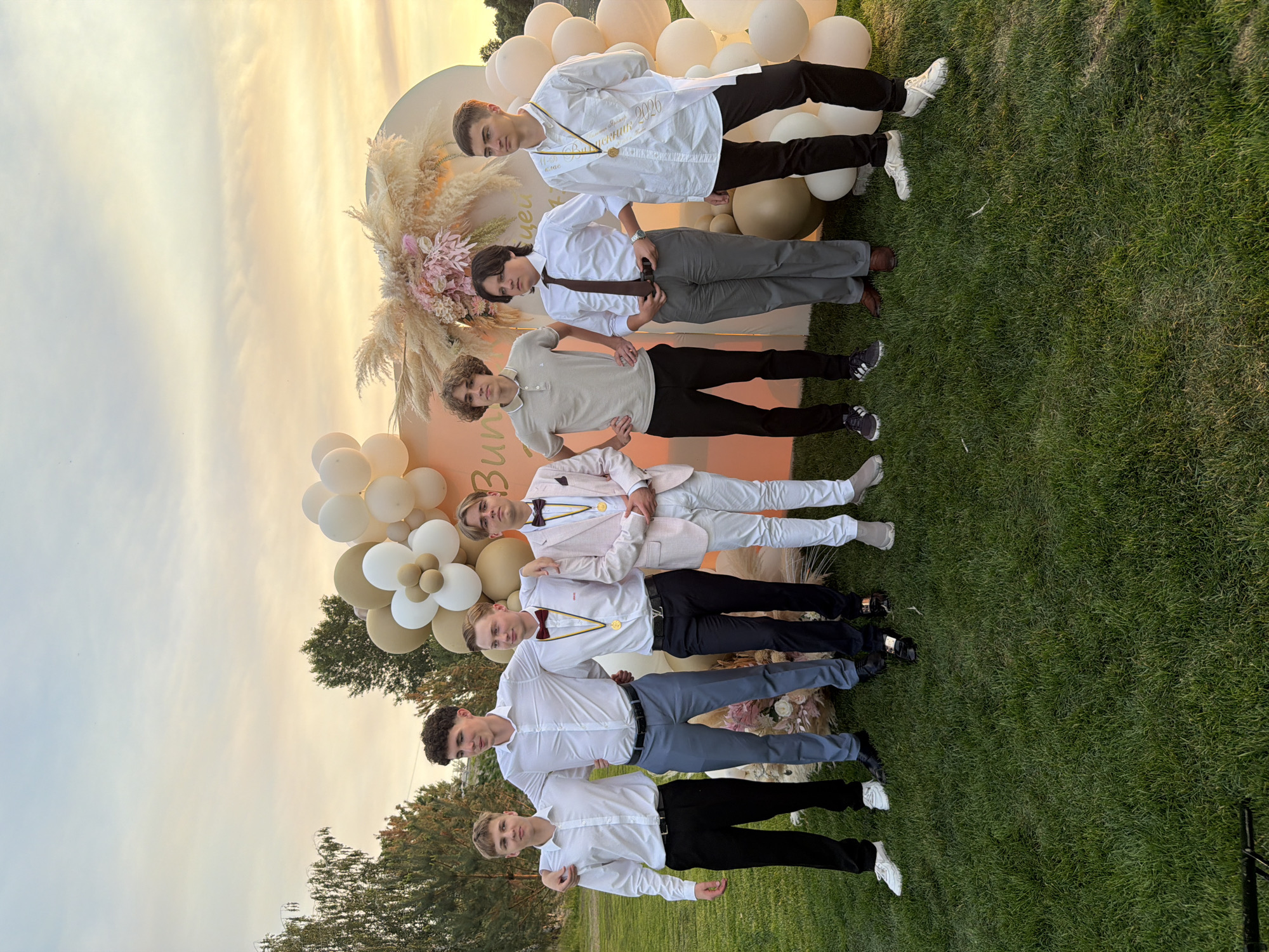 This media file depicts a group of seven young men posing for a photograph outdoors, likely at a graduation ceremony or celebratory event. The setting appears to be a grassy area with a decorative backdrop consisting of a large circular cutout, balloons in cream and gold, and dried floral arrangements. The time of day is likely late afternoon or early evening, given the warm, soft light of the setting sun illuminating the sky with hues of orange and yellow.
The subjects are all dressed formally, with several wearing white shirts and dark trousers, while others are in suits. Many of them are wearing gold medals or sashes, suggesting an award or recognition ceremony. The overall mood seems celebratory and proud.
Visible text on a white sash worn by one of the young men reads "ВИПУСК 2026," which translates to "GRADUATION 2026." This confirms the event is a graduation. The backdrop also has some faint text that is difficult to read clearly. The location is indicated as Khotianivka, Ukraine. The weather appears to be clear and pleasant.