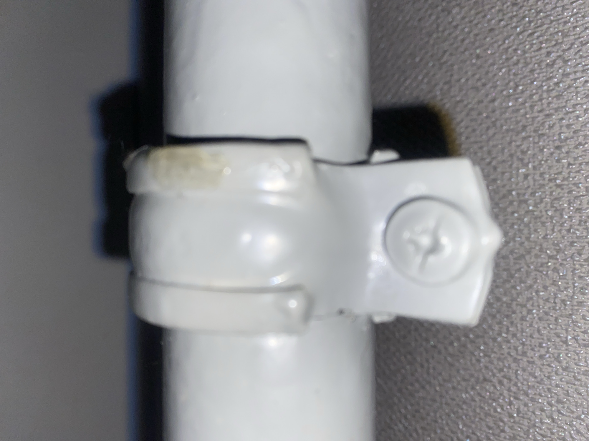 This is a close-up, forensic, matter-of-fact image of a white pipe secured by a white plastic bracket with a single screw visible in the lower portion of the image. The bracket has a split-ring design that encircles the pipe. There is a slight discoloration on the right side of the bracket. The background is blurred, showing a dark object behind the pipe and a textured surface in the foreground, possibly fabric or a textured wall. The scene is indoors, but the specific location within the city of Unknown, Unknown cannot be determined from the image.