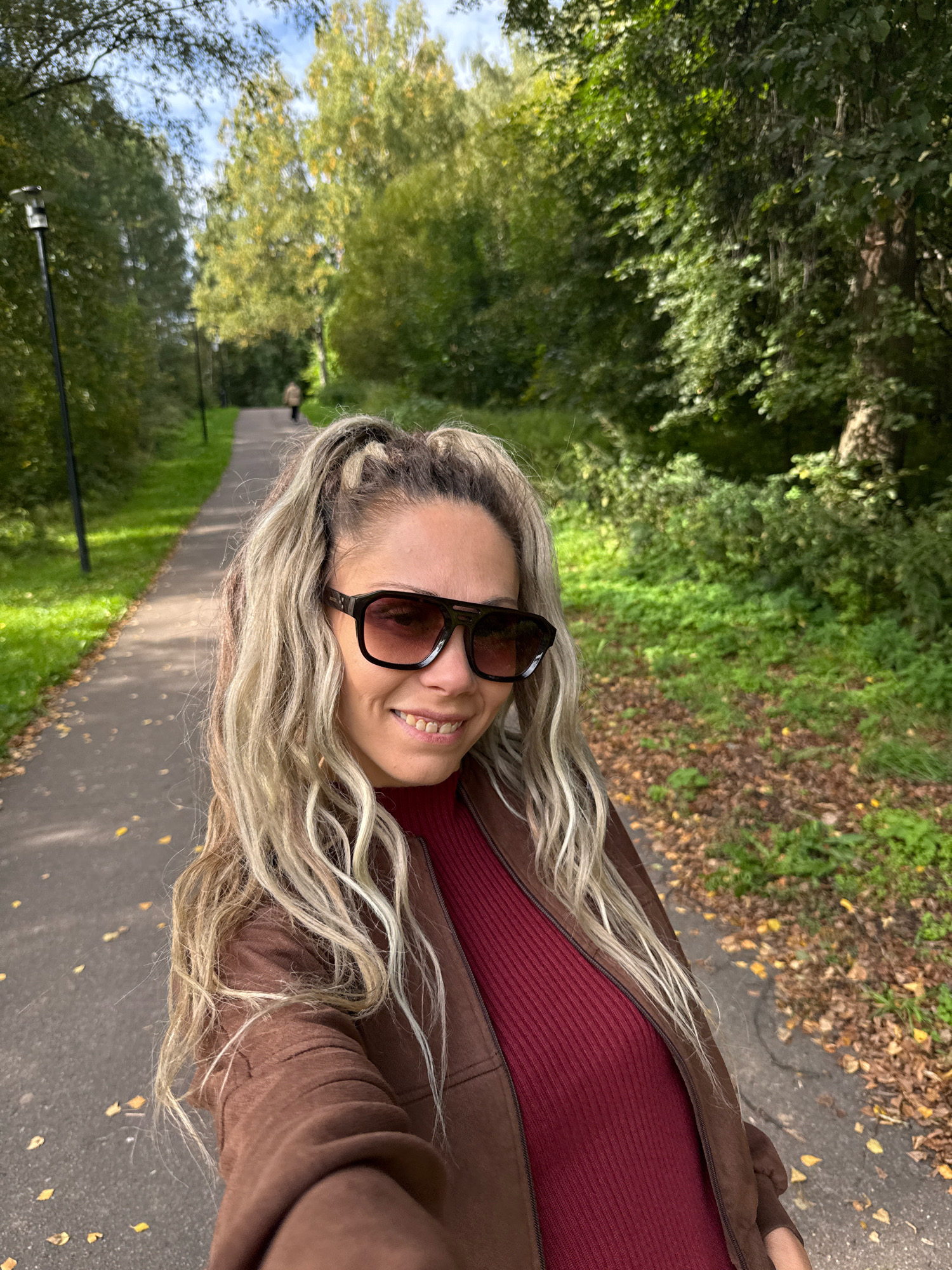 A woman with blonde hair pulled into two high ponytails and wearing sunglasses takes a selfie on a paved path in a park in Rusanovo, Russia. She is wearing a maroon turtleneck and a brown jacket. The path curves into the distance, flanked by grass and trees. Fallen leaves are visible on the ground. A single person is walking away from the camera in the background.