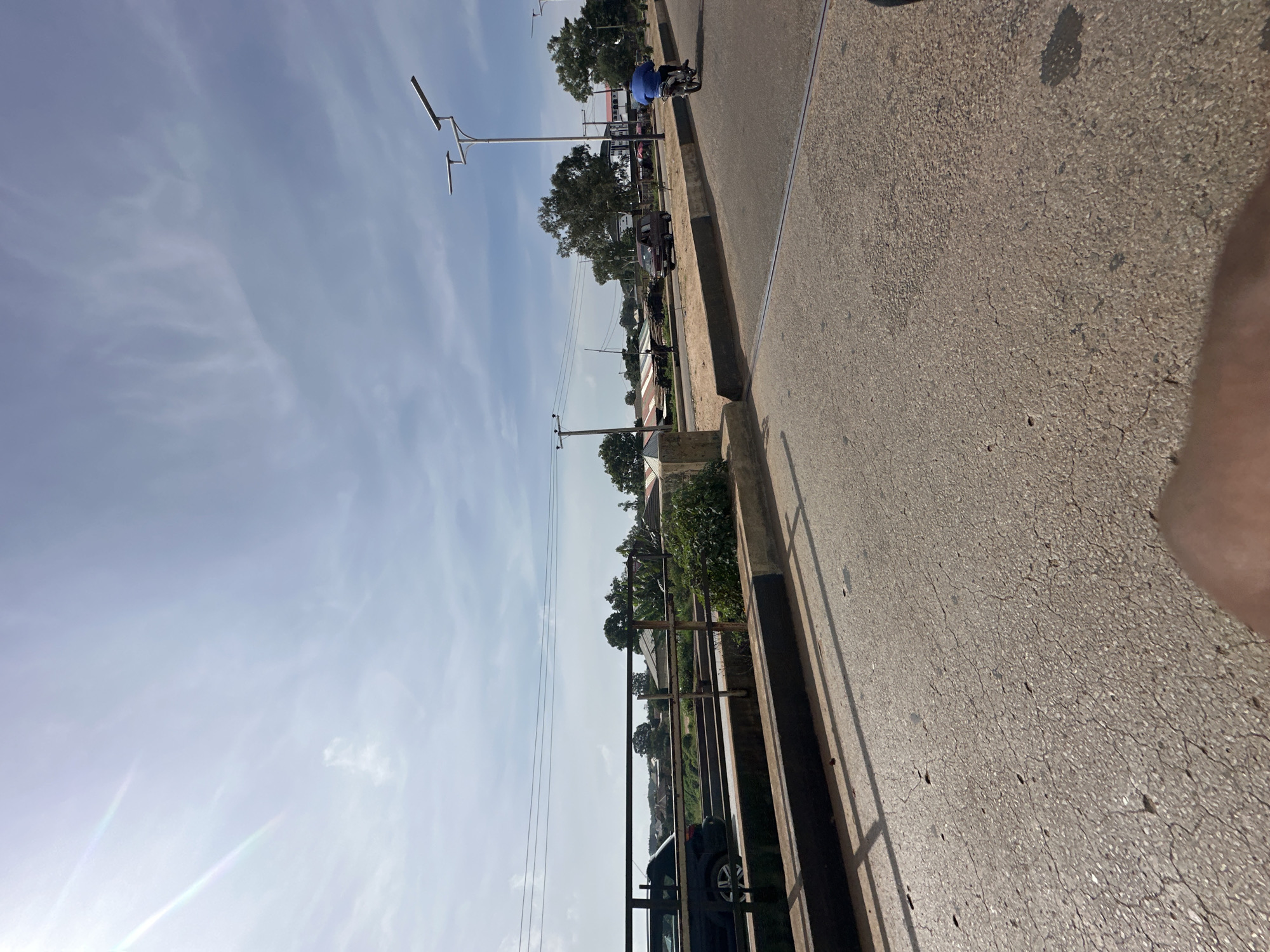 The image depicts a street scene in Bukuru, Nigeria, under a partly cloudy sky. The perspective is from a ground-level viewpoint, looking across a paved road towards a bridge and a neighborhood beyond.
On the road, a dark-colored car is parked or moving slowly on the left side, partially obscured by a railing. Further down the road, a maroon van is visible, and a person on a motorcycle, wearing a blue shirt, is riding away from the camera. Streetlights with solar panels are installed along the side of the road.
The bridge features a metal railing and a concrete structure. Beyond the bridge, residential buildings with corrugated metal roofs are scattered among trees. Power lines crisscross the sky. The road surface is asphalt with visible cracks and some debris. Shadows from the railing are cast on the road, indicating the direction of sunlight. A person's thumb is partially visible in the lower foreground, suggesting the photo was taken with a handheld device. The overall environment suggests a developing urban or semi-urban area.