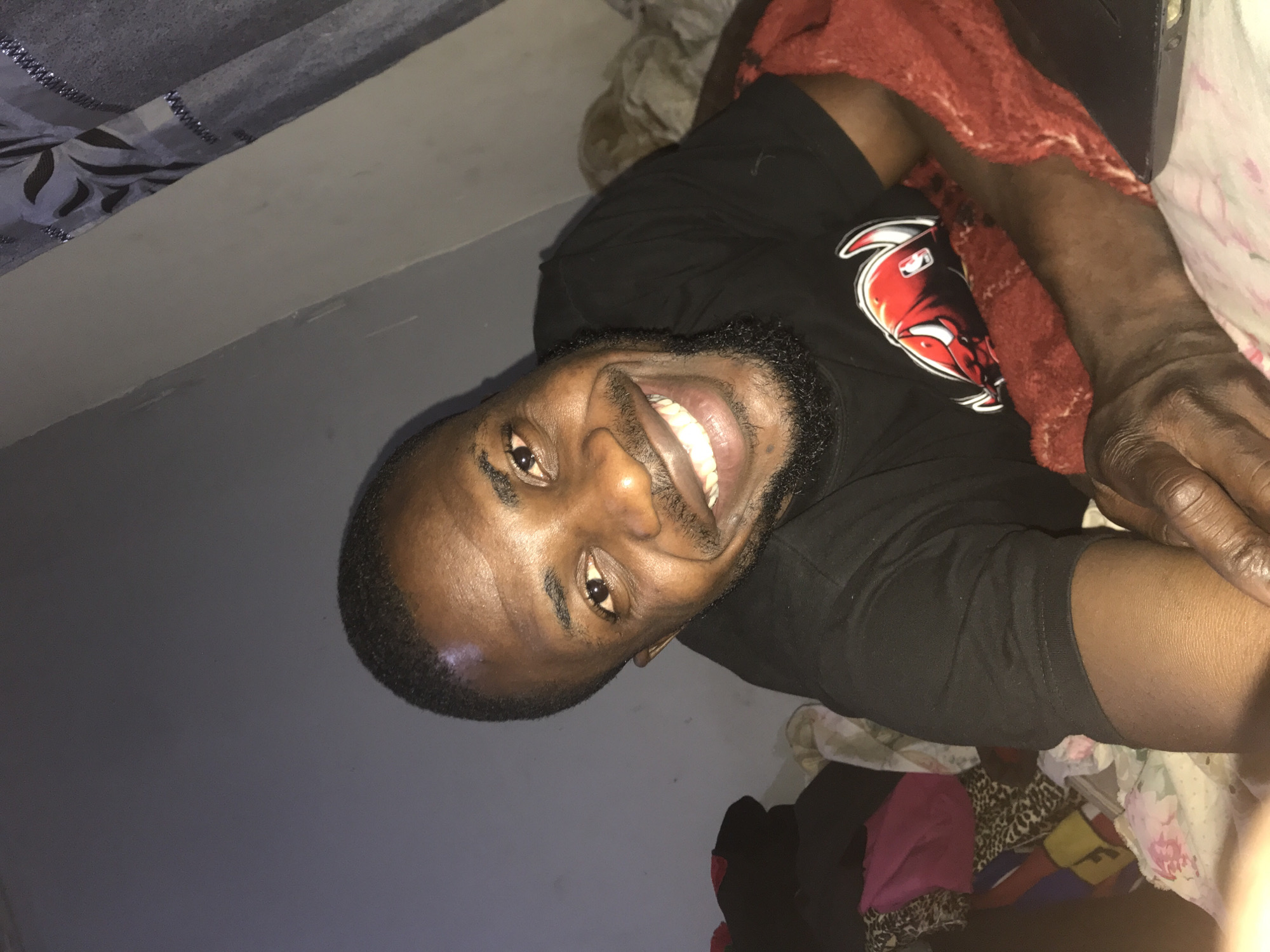 An adult male with dark skin, short dark hair, and a trimmed beard is depicted lying on a bed indoors in Rafin Par, Nigeria. He is smiling broadly, revealing white teeth, and looking directly at the camera. He wears a black short-sleeved t-shirt with a red bull head logo and 'NBA' text on the chest. His right arm rests on a red, textured blanket draped over the bed. To the left of the subject, a dark grey wall is visible, with a collection of folded textiles including dark garments, a pink bag, and a leopard-print fabric on the surface below. To the right, a grey and black patterned curtain hangs, adjacent to a lighter wall. The bedding beneath the subject is light-colored and subtly patterned.