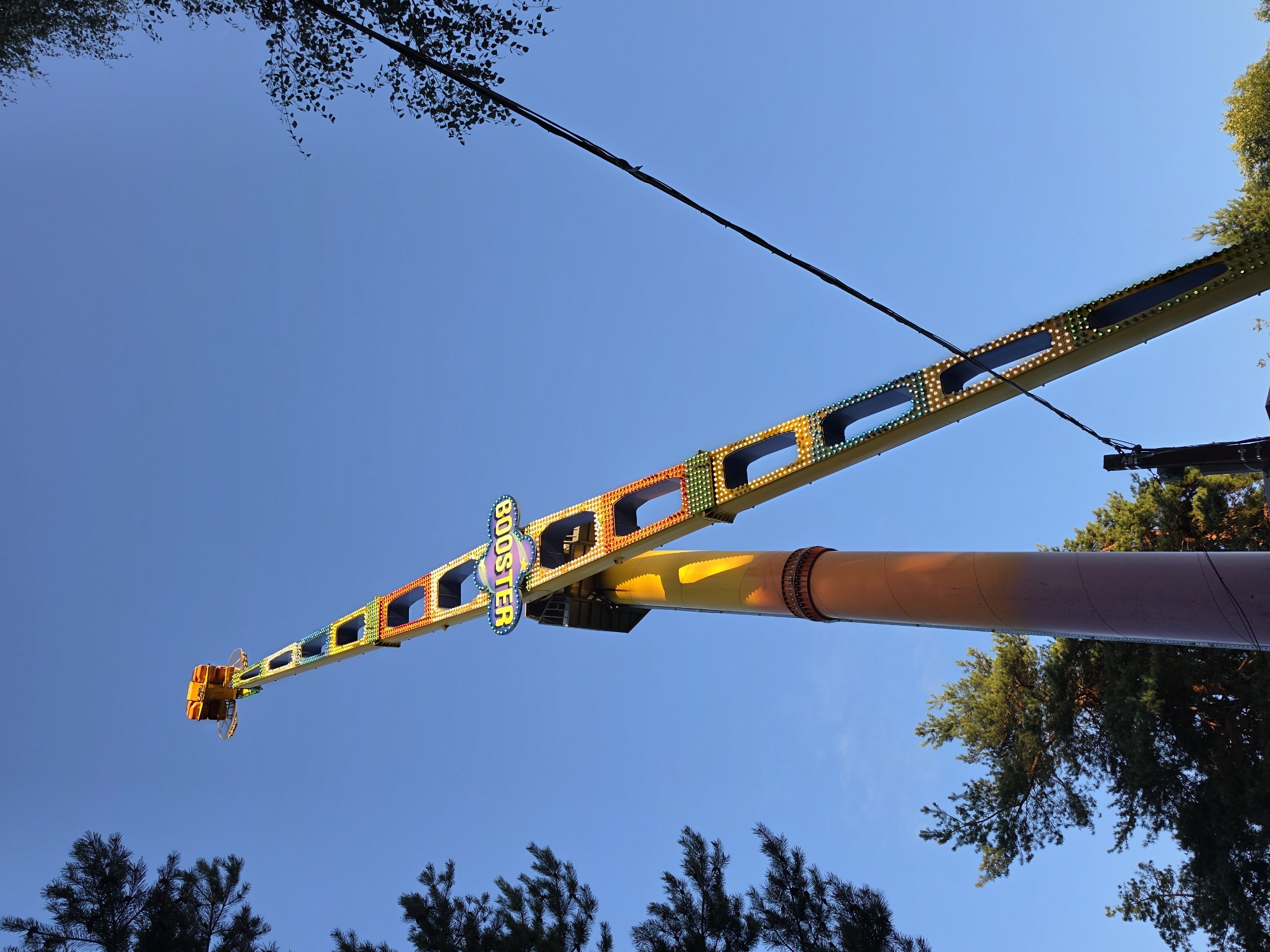 An amusement park ride called "Booster" is prominently displayed against a clear blue sky. The ride is tall and yellow, with colorful lights along its structure. The "BOOSTER" sign is visible, illuminated and attached to the ride. The base of the ride is a thick, reddish-brown cylindrical structure. In the background, there are trees, some with dark green needles and others with lighter green foliage. The scene appears to be outdoors, likely in an amusement park or fairground, with the clear sky suggesting daytime. The location is identified as Nizhny Novgorod, Russia.