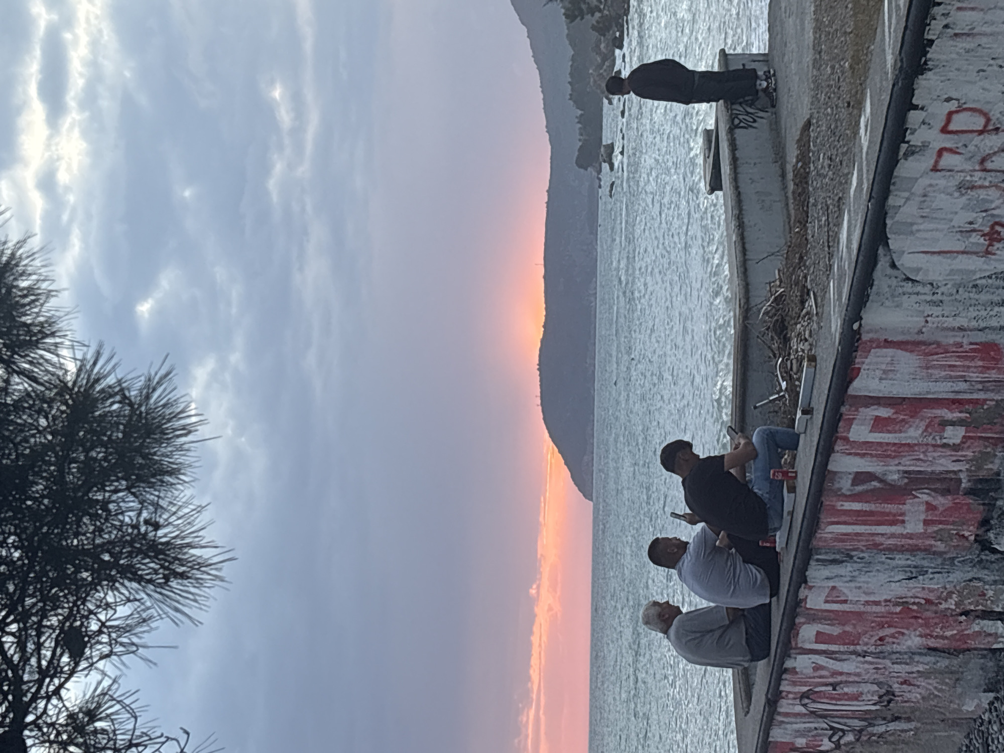 The image shows an outdoor scene at sunset. The sky transitions from blue-gray to orange-pink. A body of water extends into the horizon. A landmass is visible on the horizon. In the foreground, a concrete structure with graffiti is present. Three men are seated on this structure. One man is wearing a black shirt and blue jeans, and he is looking at his phone. The other two men are wearing gray shirts and pants. Another man is standing further along the concrete structure near the water, facing the horizon. A tree is visible on the left side of the image. The location is Bar, Montenegro.