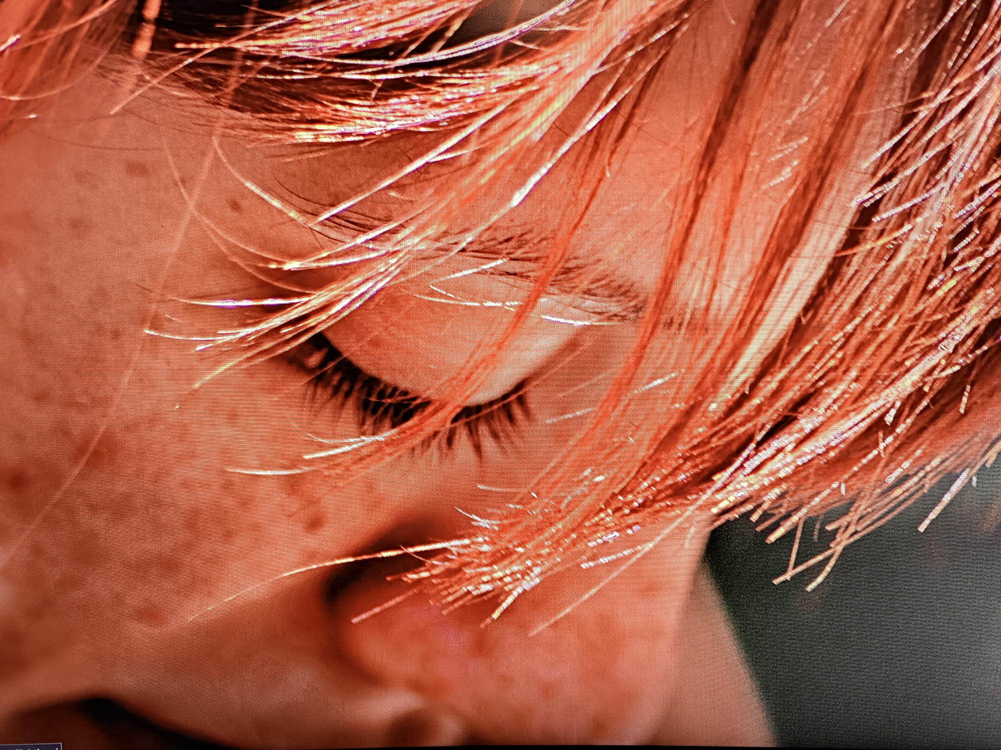 A close-up image shows a person's face, possibly a young adult or child, with fair skin and freckles visible. The person's eyes are closed or nearly closed, and eyelashes are visible. The hair appears to be reddish or light orange, with strands falling across the face. The image has an overall orange or reddish hue. The scene does not provide any context as to its location in Tbilisi, Georgia.