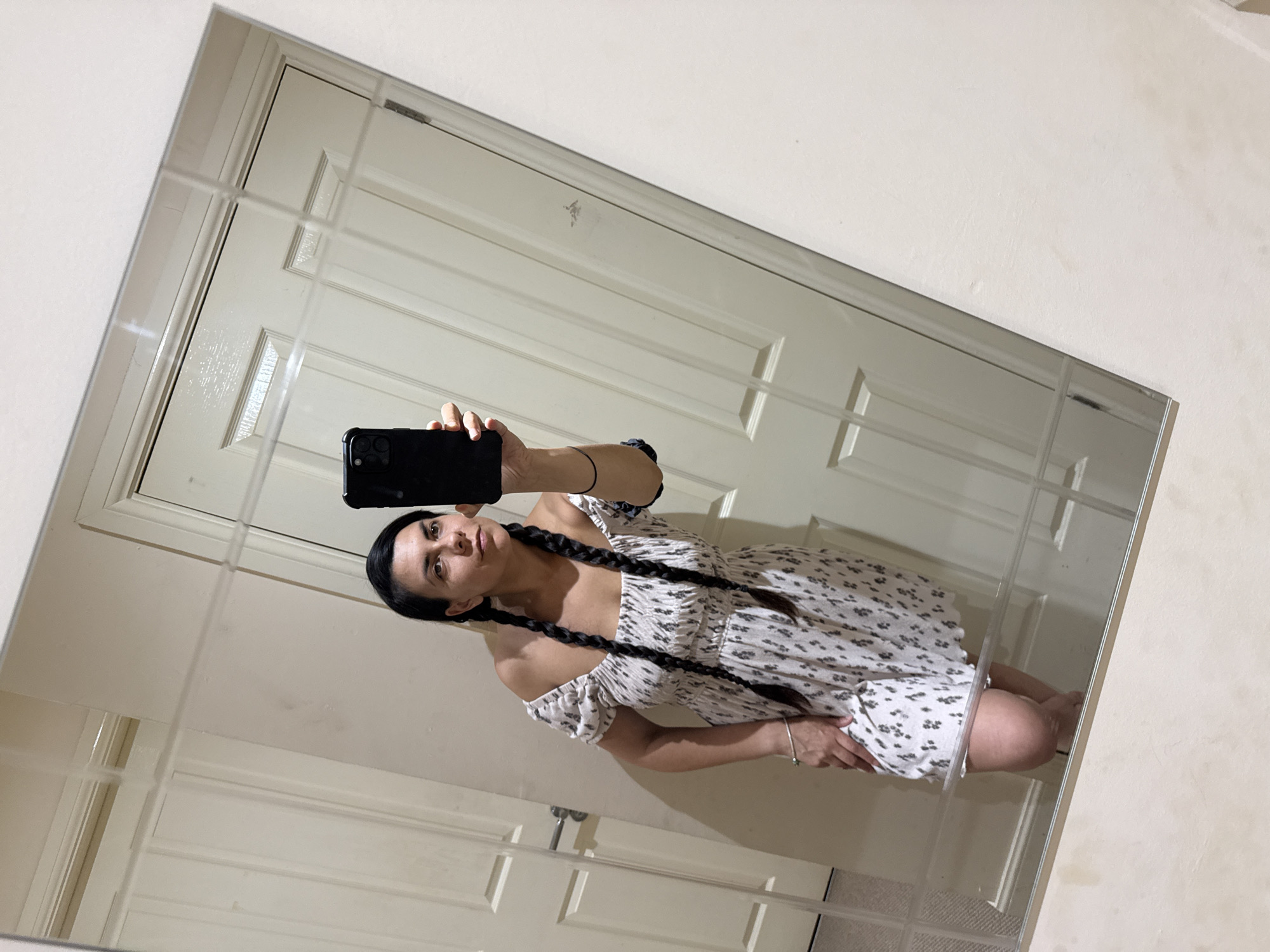 A woman with dark hair styled in two braids is reflected in a large mirror, holding up a smartphone to take a selfie. She is wearing a white off-the-shoulder dress with a black patterned print. Her left hand is on her hip, and her right hand holds the phone. The mirror reflects two white doors behind her. The woman appears to be indoors, in what could be a home environment. The location in Great Yarmouth, United Kingdom, is not specified by any visual cues in the image.