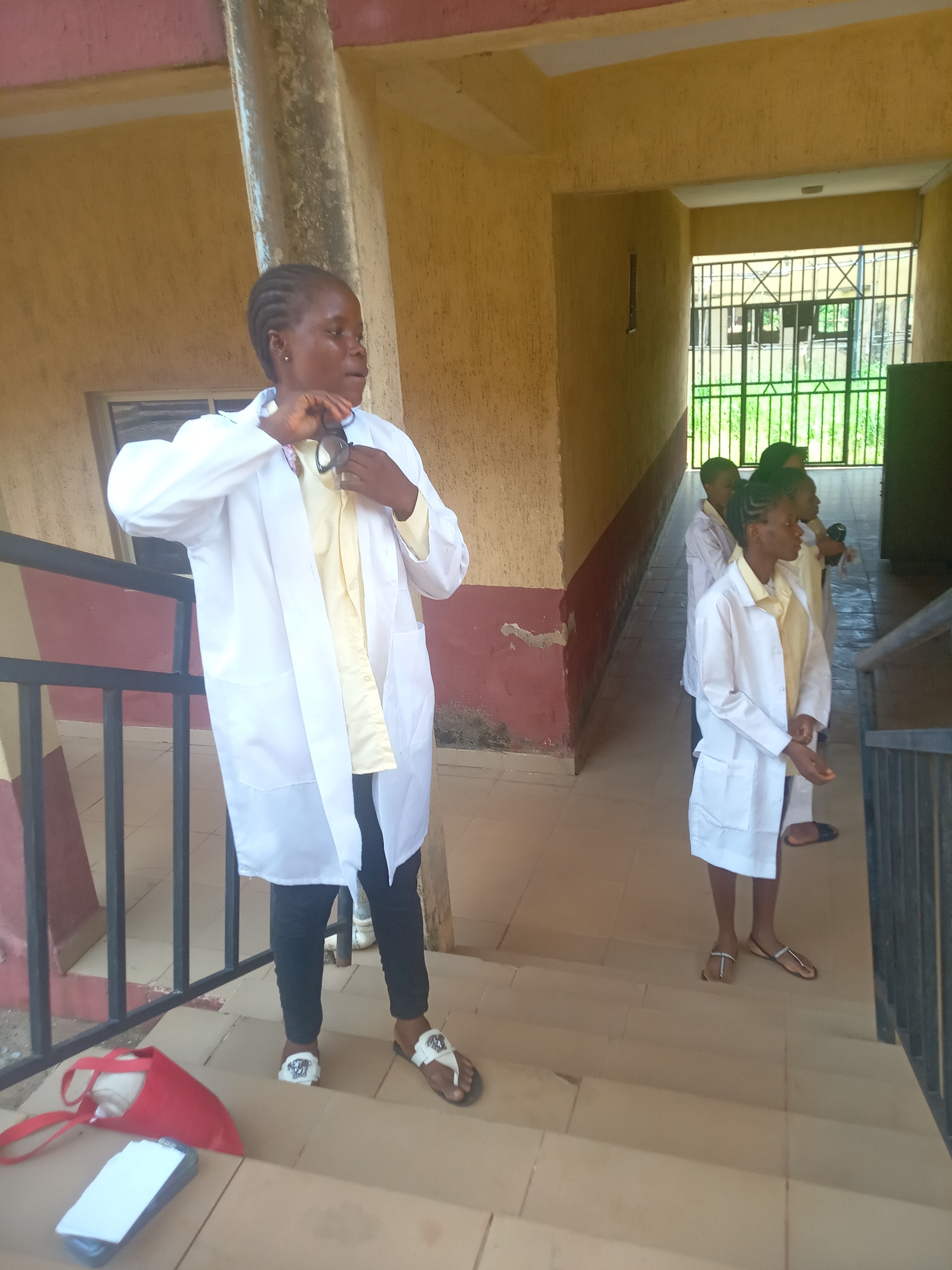 The image captures a group of young individuals, likely students or trainees, gathered in what appears to be a corridor or stairwell landing of a building in Nkwelle, Nigeria. All visible individuals are wearing white lab coats over light yellow collared shirts, suggesting an educational or practical setting related to science or healthcare.
In the foreground, a young woman with braided hair stands on a step, facing to the right. She is adjusting a pair of dark-rimmed glasses or sunglasses with both hands. She wears black trousers and white flip-flops with a prominent metallic emblem on them. Beside her feet on a lower step, a red fabric bag and a white notebook or clipboard resting on a dark rectangular object are visible.
Further down the corridor, to the right and slightly obscured, are at least three other individuals also dressed in white lab coats and light yellow shirts. They appear to be standing on steps or in a line, looking forward or slightly to the side, possibly waiting.
The setting features light brown/tan colored walls, with a reddish-brown lower section on the left. Black metal railings run alongside the steps. In the background, a large metal gate with a grid pattern can be seen, beyond which green foliage is visible, indicating an outdoor area. The natural light suggests it is daytime. The overall atmosphere appears neutral, with the individuals seeming to be in a moment of transition or waiting.