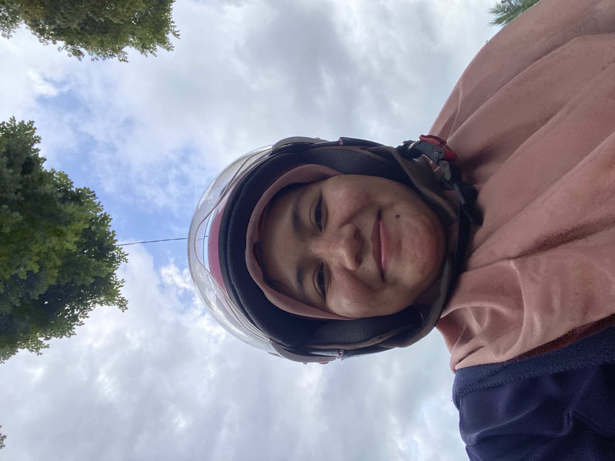 A woman wearing a pink hijab and a motorcycle helmet is captured in a close-up, low-angle selfie. She is looking at the camera with a smile. The background consists of a cloudy sky with glimpses of green foliage from trees. The image appears to have been taken outdoors, likely in a location with natural surroundings. The woman's attire suggests she is either traveling by motorcycle or has recently dismounted. The overcast sky indicates potentially moderate weather conditions.