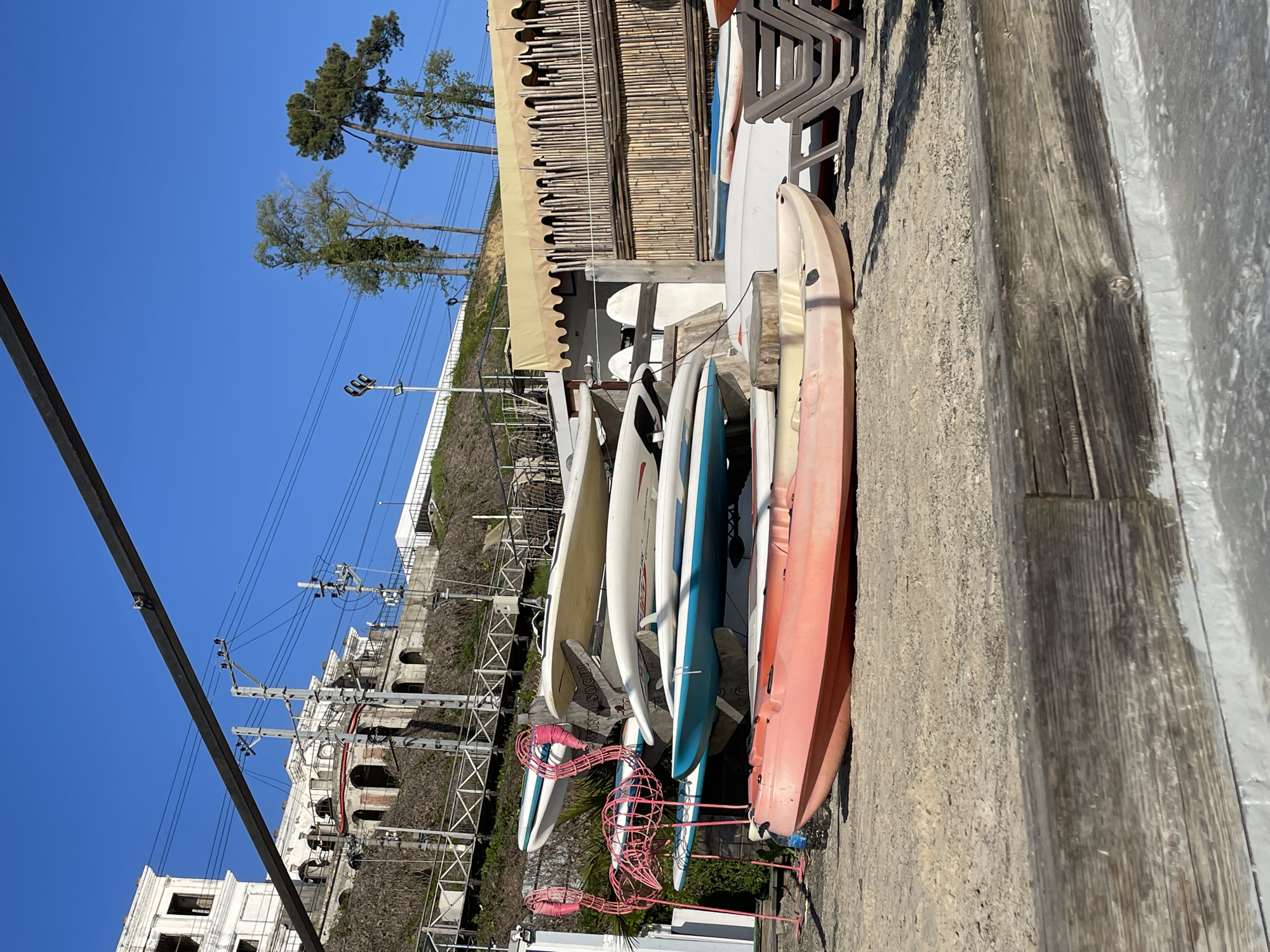 The image captures a scene on the coastline of Sochi, Russia. Various surfboards and kayaks are lined up against a weathered wooden barrier, presumably for rent or sale. A pink, metal flamingo sculpture stands among the surfboards, adding a touch of whimsy. In the background, a building with arched windows and a light-colored facade can be seen, partially obscured by foliage. Above the building, utility poles and power lines stretch across a clear blue sky. Further back, a few tall trees are visible. The scene is captured during daylight hours with bright sunlight illuminating the objects and structures. A building with a bamboo facade is partially visible in the upper right portion of the scene.