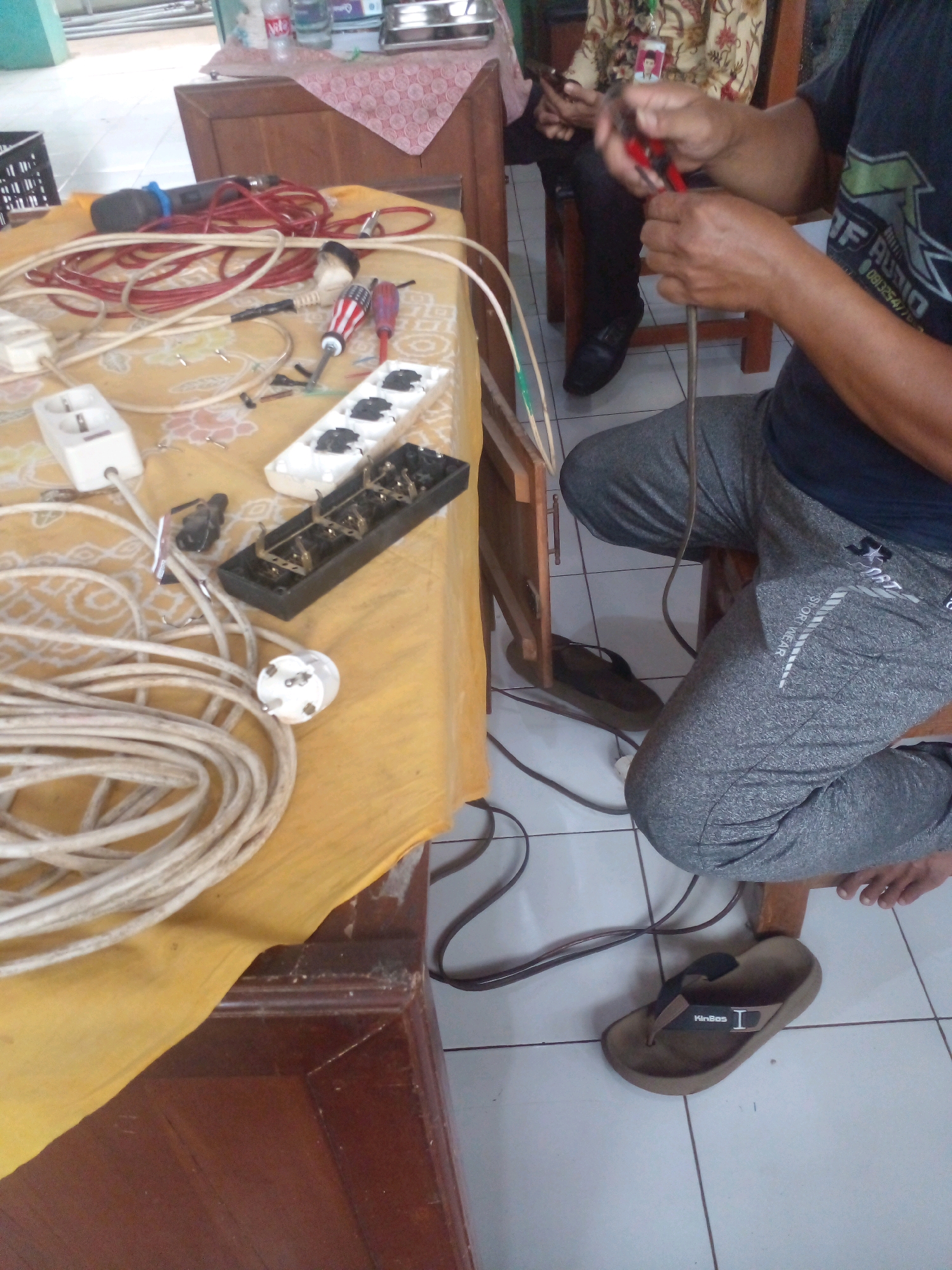 An overhead medium shot captures a scene on a table and the floor in what appears to be an indoor setting with tiled flooring. The table, covered with a yellow cloth, is laden with electrical equipment. Prominently featured are various power strips, extension cords, a microphone, screwdrivers, and electrical plugs and sockets.
In the foreground, a person, partially visible, is seated and engaged in using a pair of red-handled wire cutters to strip a piece of wire. The person is wearing a dark t-shirt with white lettering and grey sweatpants. A brown flip-flop is visible on the tiled floor to the right of the person.
In the background, another individual is seated, wearing a batik-patterned shirt and holding a mobile phone. The environment suggests a workshop or a space where electrical repairs or modifications are being conducted. The overall impression is one of practical work being undertaken.