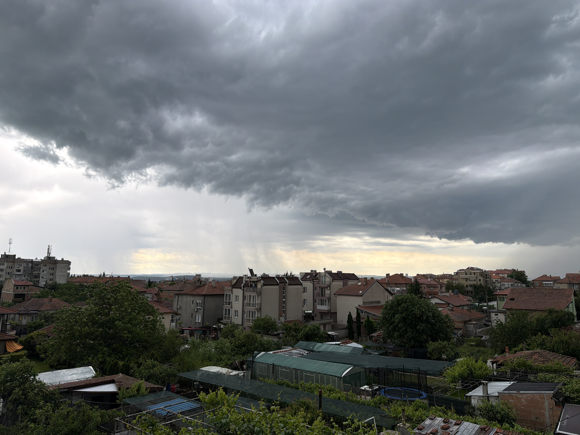 The image depicts an overcast sky over Burgas, Bulgaria. Dark storm clouds dominate the upper half of the scene, with streaks of rain visible in the distance. The lower half showcases a residential area with buildings of varying heights and styles, interspersed with trees and greenery. Some greenhouses and gardens are visible in the foreground. The building rooftops are primarily red tile. The general atmosphere is dark and impending rain.