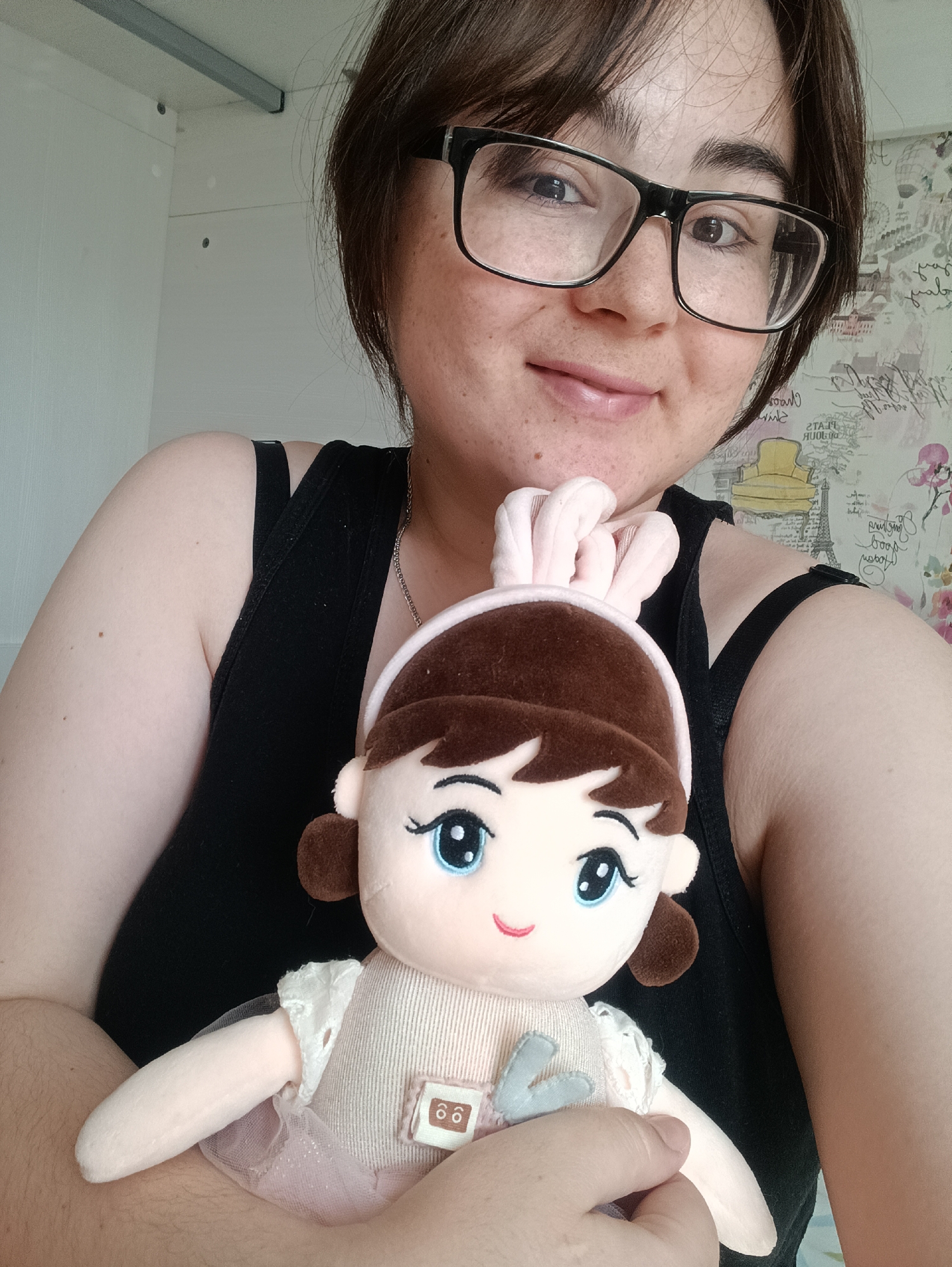 A person with dark hair and glasses is holding a plush doll with bunny ears. The person is wearing a black tank top. The background includes a white wall and patterned wallpaper. The location is Unknown, Unknown.