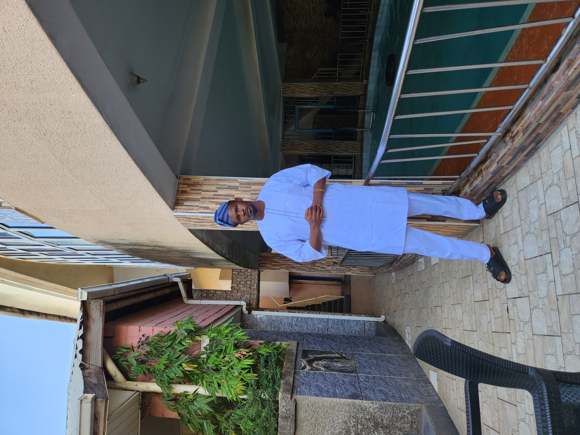 A man is standing on a tiled surface adjacent to a structure with a green and orange awning. He is wearing a white outfit with a blue cap and black sandals. The location appears to be an outdoor area, possibly a patio or walkway, with a view into a passageway with stairs. Lush greenery is visible next to a building with pink brickwork. A black chair is partially visible on the tiled surface. The city and region are Unknown, Unknown.