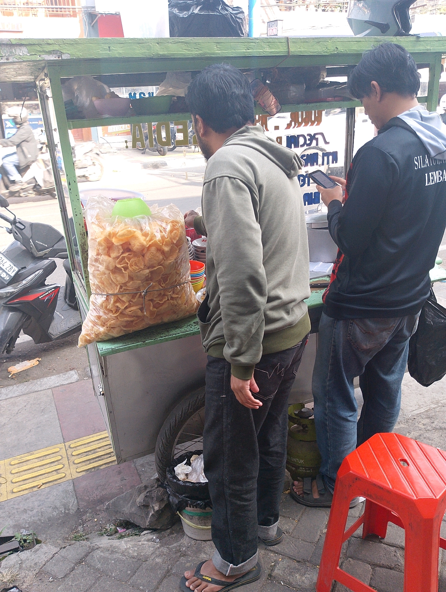 In Lembang, Indonesia, two young men are observed at a street food cart. The cart, painted green, is laden with food items. A large, clear plastic bag overflowing with fried snacks occupies a prominent position on the cart. To the left, a motorcycle is parked, its front visible. A person in a grey hoodie stands with their back to the viewer, facing the cart. Another person, wearing a black long-sleeved shirt and blue jeans, is engrossed in their mobile phone, also facing the cart. In the foreground, a bright red plastic stool stands empty. The ground is paved with stone and concrete, with a tactile paving strip to the left. A gas cylinder is visible behind the cart. The overall scene depicts a typical street-side commerce interaction in Indonesia.