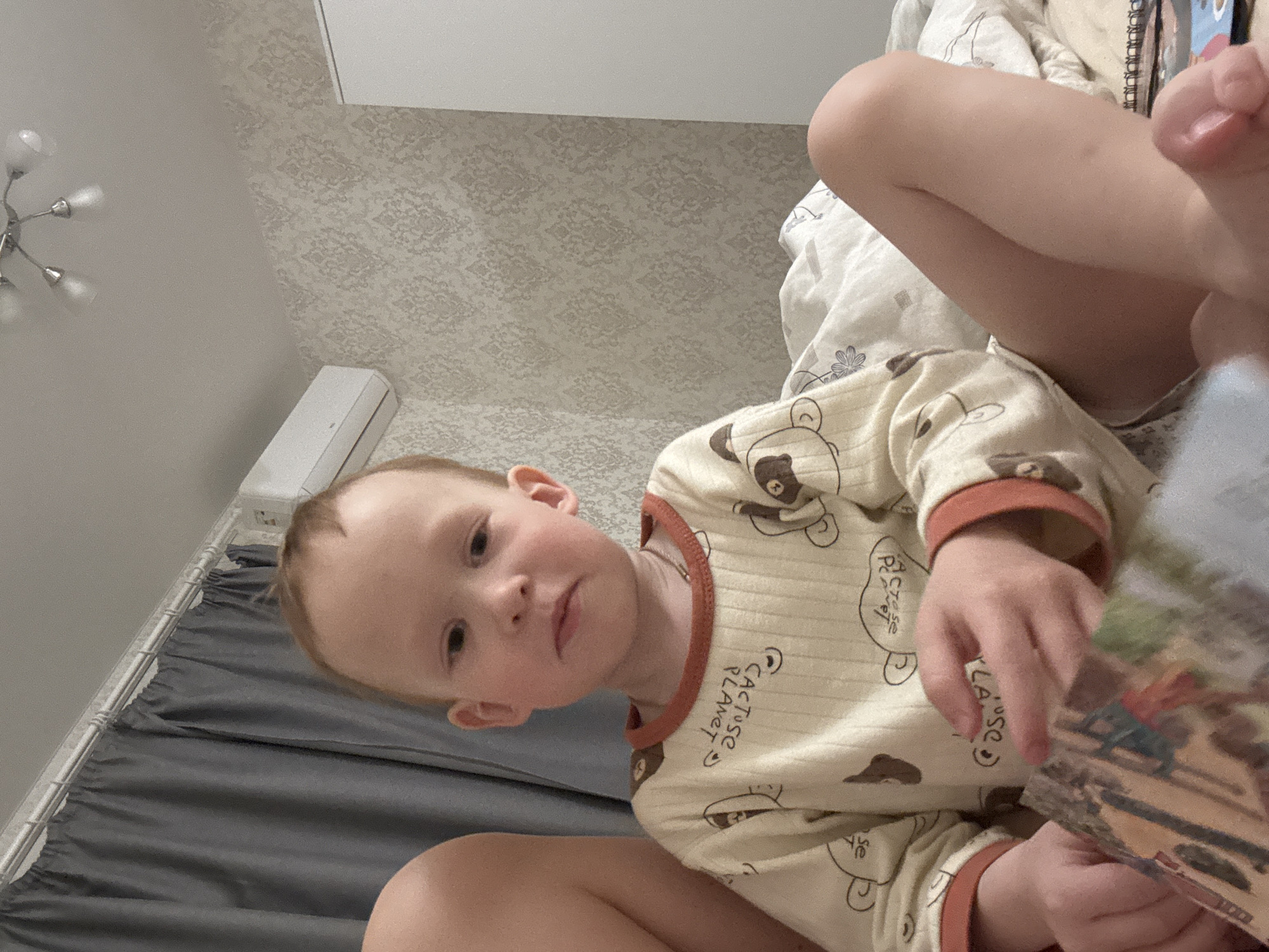 A young child, presumably in a residence located in the city of Ферма, Russia, is seated on a bed. The child is fair-skinned with reddish-blonde hair. The child is wearing light-colored pajamas with a bear design and brown trim around the neck and wrists. The pajamas have the phrase "Cactus Planet" printed on them. The child is looking at the camera with a neutral expression. The bed has grey sheets and a white patterned comforter. Part of another person is visible in the upper right of the frame, including a leg and foot. A book or magazine is partially visible in the child's hands. The room's wallpaper is patterned, and a light fixture is visible in the upper left corner. An air conditioning unit is visible on the wall.