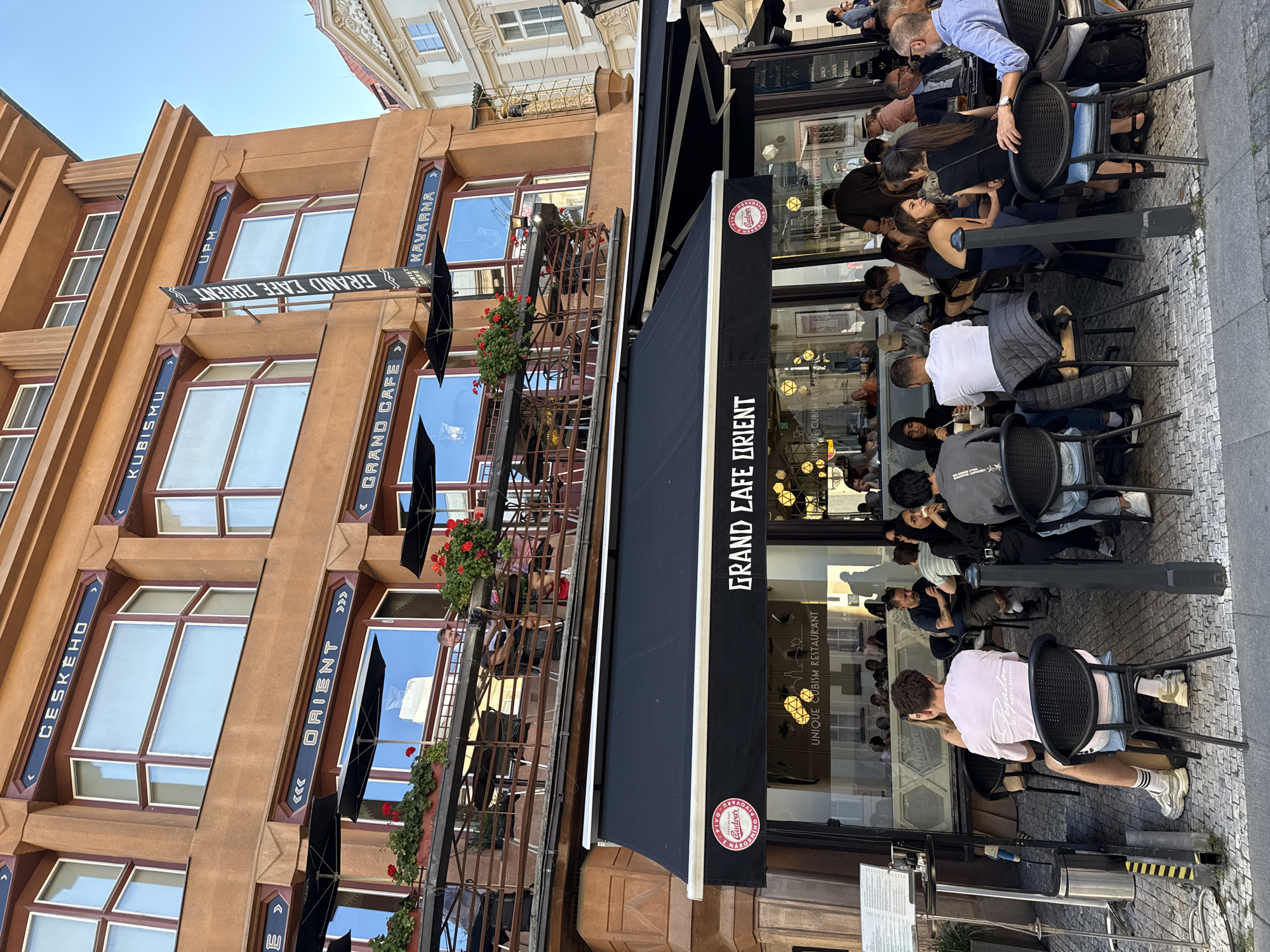 This is a photograph of the Grand Cafe Orient, a historic establishment located in Prague, Czechia, specifically on the second floor of the House of the Black Madonna. The cafe's black awning is prominently displayed, featuring the name "GRAND CAFE ORIENT" in white lettering. Below the awning, through the large glass windows, patrons can be seen seated at tables. Some of these tables are also set up outside on the sidewalk, where people are dining and socializing.
The building itself is an example of Cubist architecture, characterized by its angular and geometric facade, painted in a warm, terracotta orange color. Decorative elements in dark blue with gold accents are visible on the exterior, including signage that reads "ČESKÉHO," "KUBISMU," "UPM," "ORIENT," and "KAVÁRNA." Red geraniums bloom in planters along the balconies, adding a touch of color to the scene. The sky above is a clear blue, indicating it is likely daytime. The overall impression is a lively outdoor dining scene in a culturally significant architectural setting.
