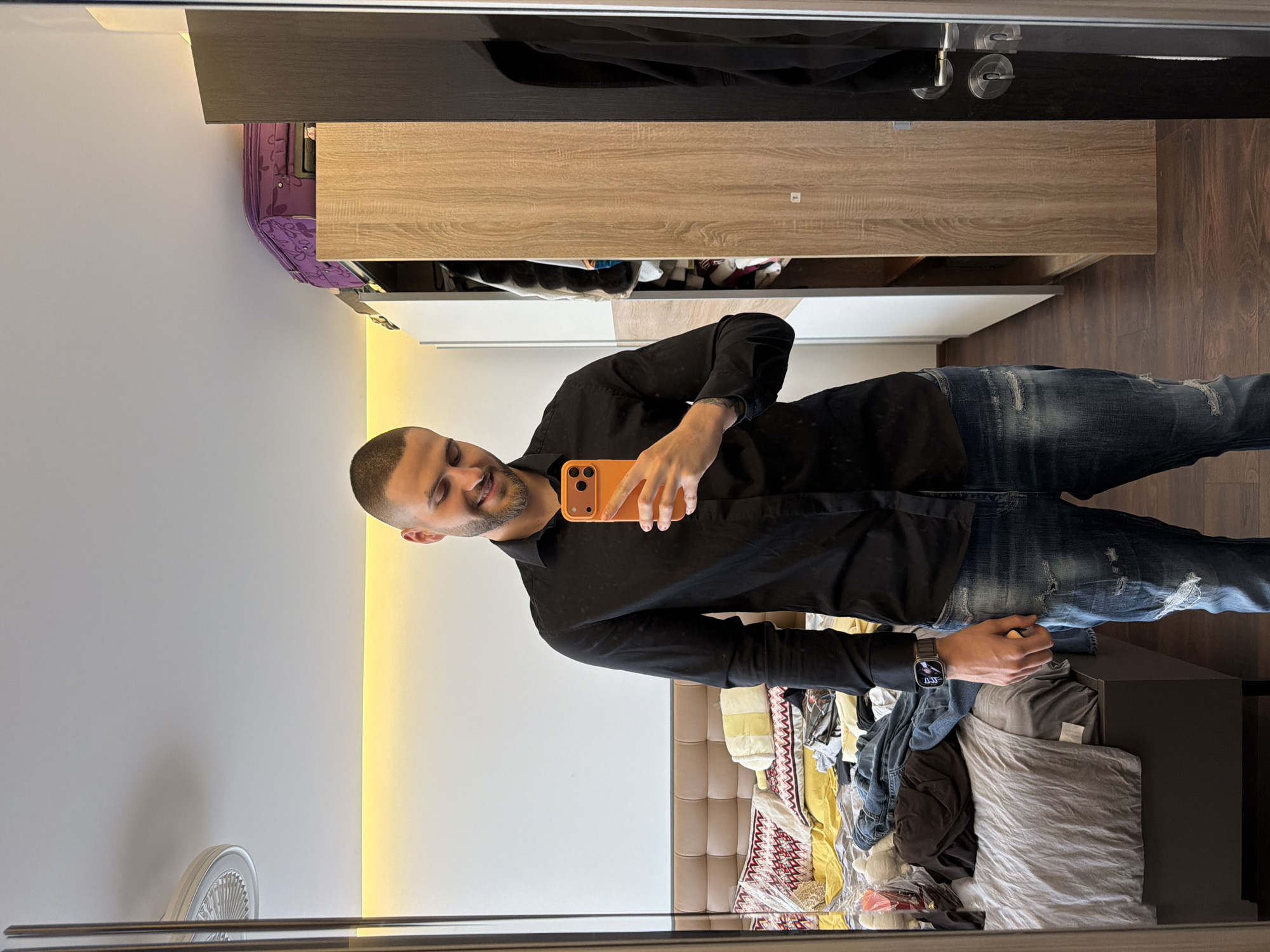 A man is taking a selfie in a mirror in a room in Sofia, Bulgaria. He has short dark hair and a beard and is wearing a black button-down shirt and blue jeans with rips. He is holding an orange smartphone. Behind him is a bed with several piles of clothes on it, including a white comforter with a red and black pattern. To the left of the man is a white wall with a strip of light. To the right of the man, there is a dark gray dresser, and behind the man is a wardrobe with light wood grain. There is a purple suitcase on top of the wardrobe. The room appears to be messy.