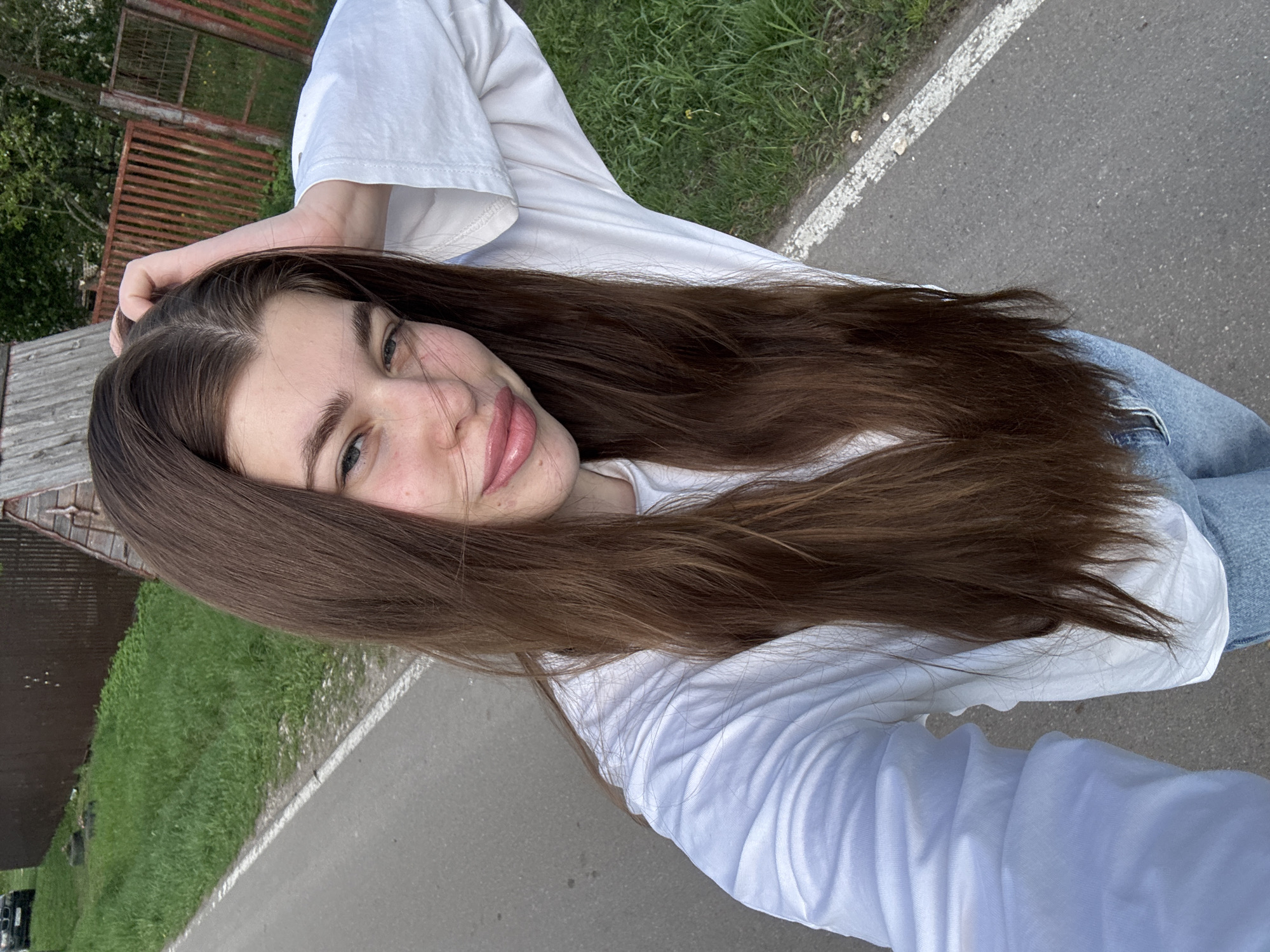 A woman with long, brown hair is the primary subject of the photograph. She is wearing a white long-sleeved shirt and light blue jeans. She is holding her hand near her head, seemingly adjusting her hair. The setting appears to be outdoors on a paved path next to a grassy area. In the background, there's what appears to be a wooden structure or building, and a red metal fence. The environment seems to be a suburban or park-like area, although the location in the city of Meshchery, Russia, cannot be definitively confirmed from the visual data alone.