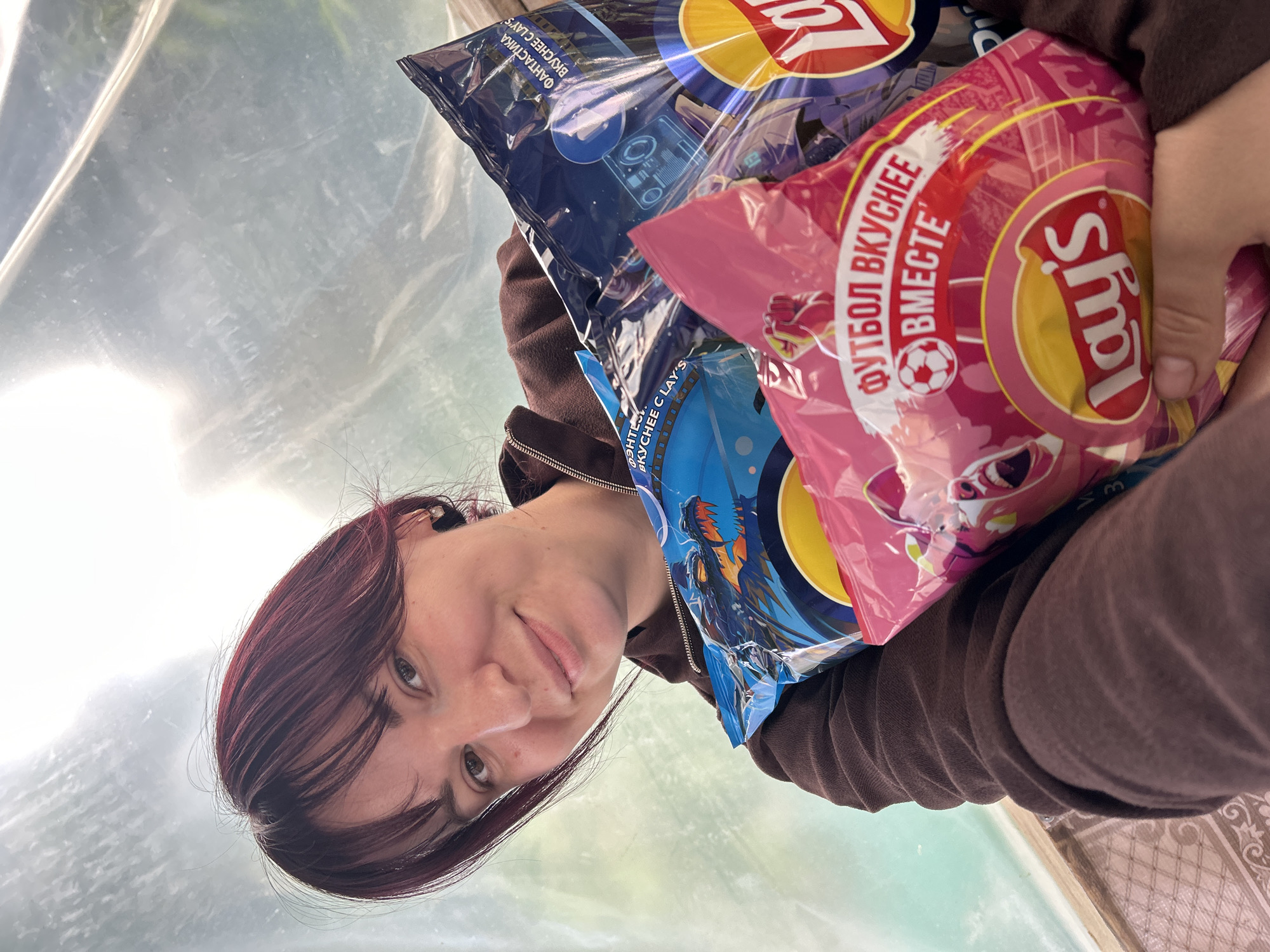 A young adult female with dark reddish-brown hair and bangs smiles directly at the viewer while holding a stack of three Lay's potato chip bags. She is wearing a dark brown hooded zip-up sweatshirt. The bags are prominently displayed; a pink bag features the Russian text "ФУТБОЛ ВКУСНЕЕ ВМЕСТЕ" (Football is tastier together) and a soccer ball icon, a blue bag displays a dragon illustration and "ФЭНТЕЗИ ВКУСНЕЕ С LAY'S" (Fantasy is tastier with Lay's), and a dark blue/purple bag shows sci-fi graphics and "ФАНТАСТИКА ВКУСНЕЕ С LAY'S" (Science Fiction is tastier with Lay's). The background is a brightly lit, diffuse area enclosed by translucent plastic sheeting, suggesting a greenhouse or covered outdoor space. A wooden frame and a patterned ground surface are visible in the lower left corner. The scene is located in the city of Фоки, Russia.