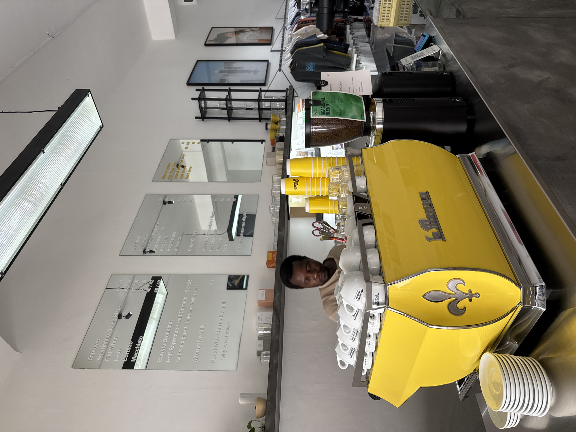 The scene depicts the interior of a cafe, likely in Cape Town, South Africa, focusing on the barista station. A man is partially visible behind a bright yellow espresso machine, possibly interacting with it. Cups and saucers are neatly stacked on the machine, alongside yellow paper cups and various tools. The coffee beans are in a grinder. The menu, detailing espresso-based drinks and alternative milk options, is mirrored on the wall. Framed pictures and shelving are visible higher up on the wall.