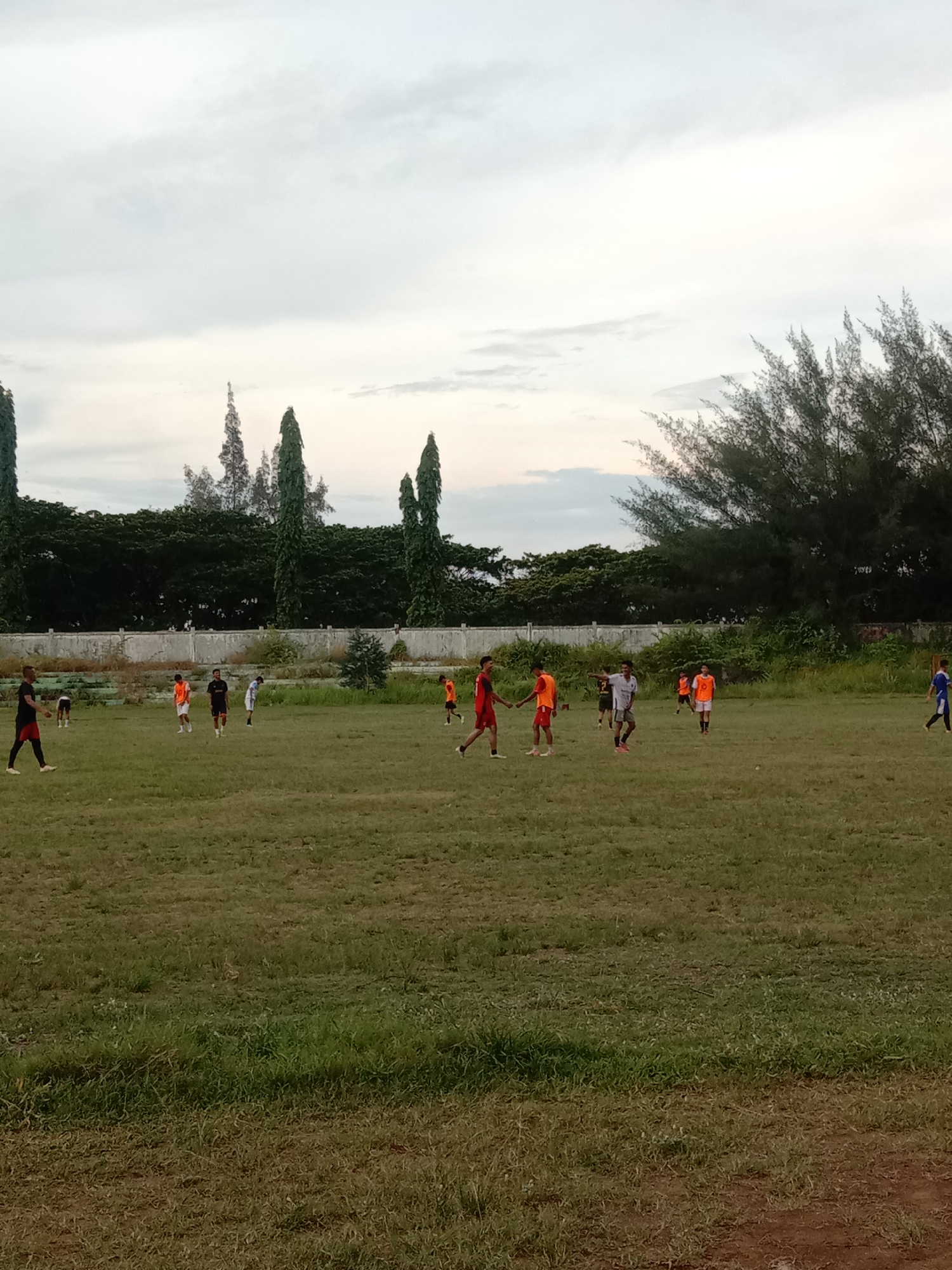 The image depicts a soccer practice taking place on a grassy field in Lhokseumawe, Indonesia. A group of young men are scattered across the field, some wearing orange jerseys, others in darker colors. Trees and a low wall form the background of the scene. The sky is overcast.