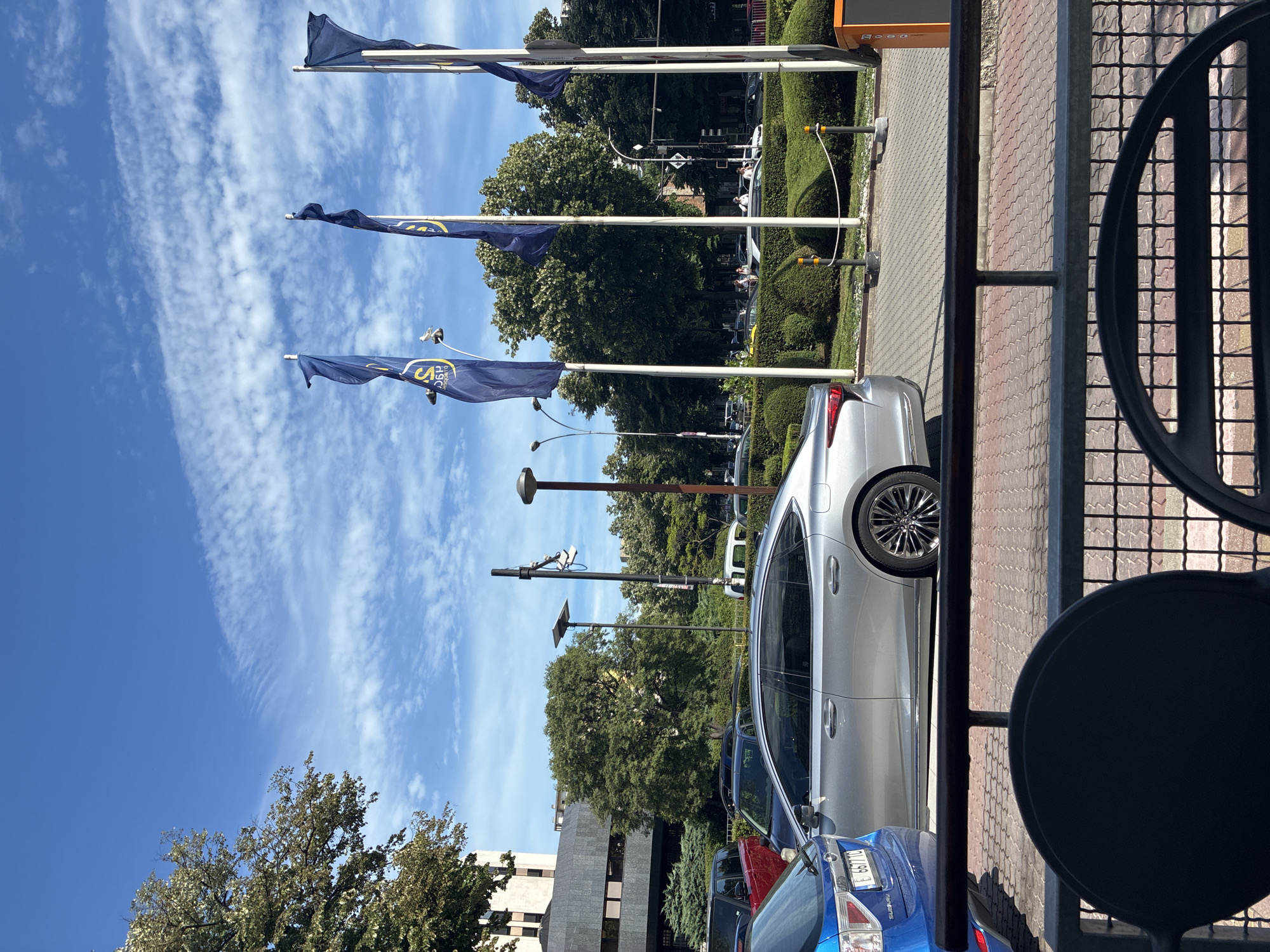 The image depicts an outdoor scene with flags, cars, and greenery. Three dark blue flags with a yellow logo are prominently displayed, fluttering in the wind. The flags appear to be on tall poles. In the foreground, the side of a silver car is visible, parked on a paved area. Behind the car, a row of other vehicles, including a blue Subaru, is parked. A building with a dark facade and several windows is partially visible on the left side of the image.
The background features lush green trees and manicured bushes, suggesting a well-maintained outdoor space, possibly a park or a public area. The sky is blue with scattered white clouds, indicating fair weather. The lighting suggests it is daytime, likely midday or afternoon. The scene appears to be a street or parking area with some commercial or civic buildings nearby. There are lampposts and possibly some street furniture or infrastructure visible.
The image is captured in Varna, Bulgaria. No specific activity or event is clearly taking place, but the presence of parked cars and flags suggests a public or commercial setting. The overall mood is calm and bright. Visible text on the blue Subaru's license plate is "B 667 TC". The logo on the flags is yellow and appears to be circular with some lettering within it, though the text is not legible.