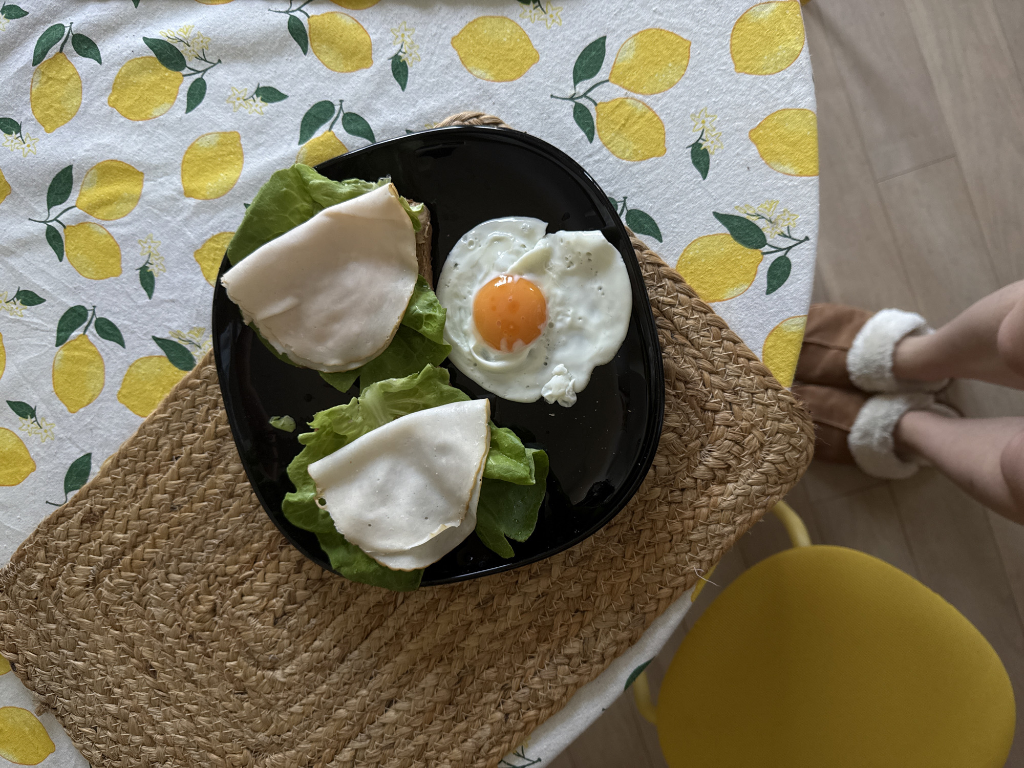 The image shows a black plate on a woven placemat, containing two open-faced sandwiches with lettuce and sliced turkey breast, and a fried egg. The placemat rests on a table with a lemon-patterned tablecloth. A yellow chair is partially visible in the lower right corner. Part of a person's legs wearing slippers are visible in the upper right corner. The location is Vozdovac Urban Municipality, Serbia.