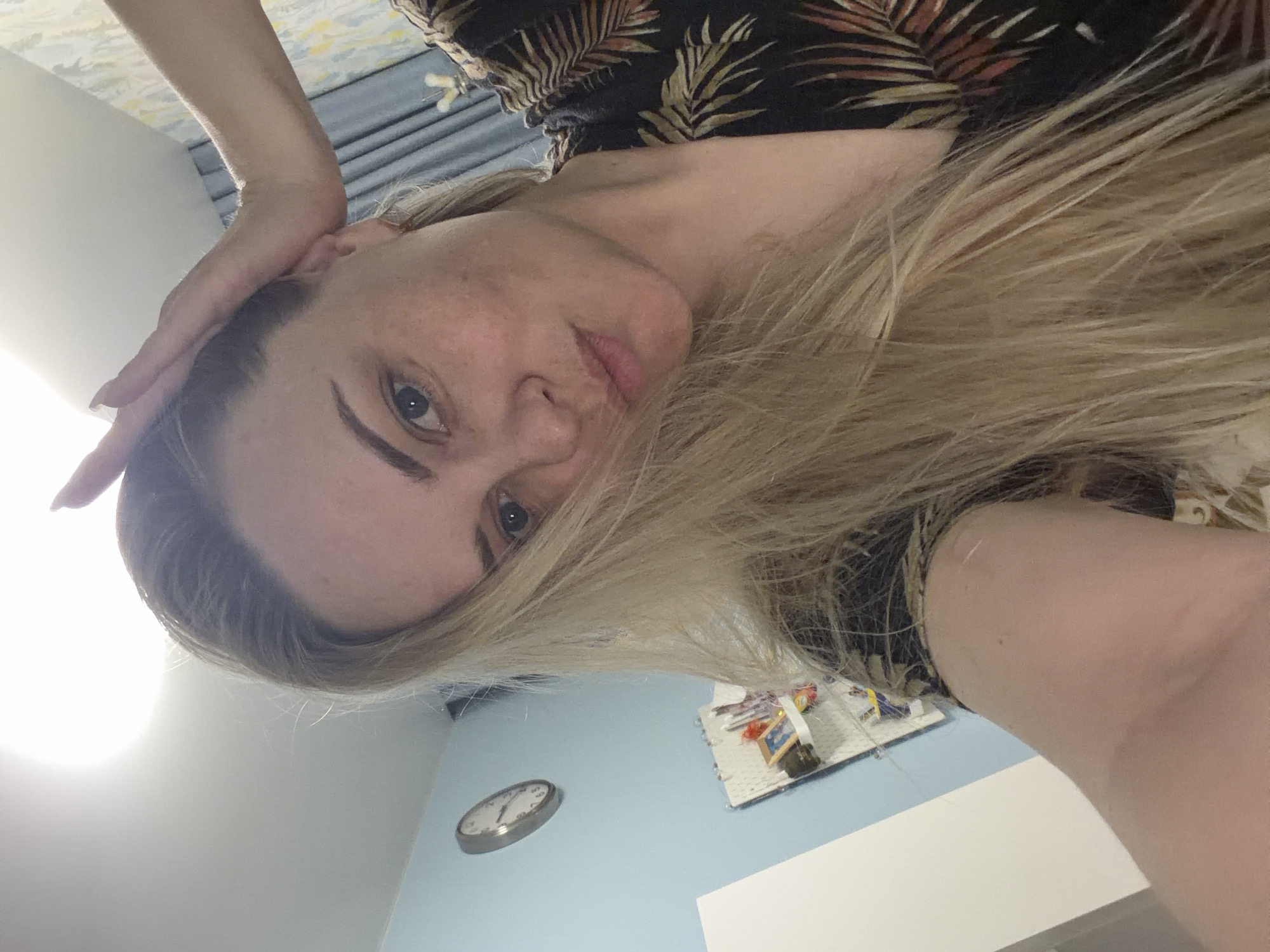 A woman with blonde hair is looking down at the camera. She is wearing a dark, tropical-print shirt. She has her hand on her head. Behind her, a blue wall is visible. A clock and a bulletin board with various items are on the wall.
It is impossible to determine the location in the city of Votkinsk, Russia, from this image.