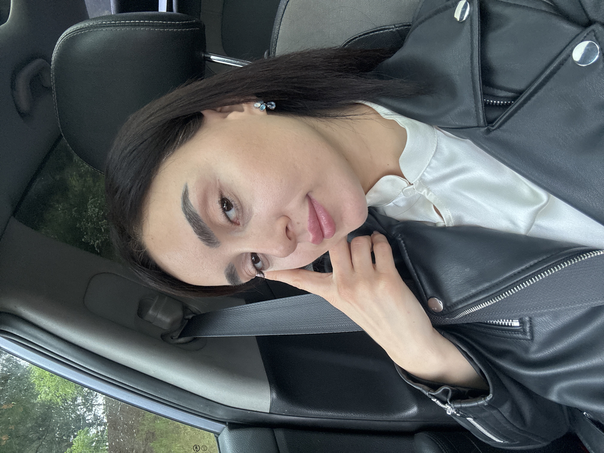 A woman is taking a selfie inside a car. She is wearing a black leather jacket over a white blouse. She is wearing earrings. A seatbelt is visible. The car is likely parked, with trees visible through the window. Location details are not discernible to confirm the city.
