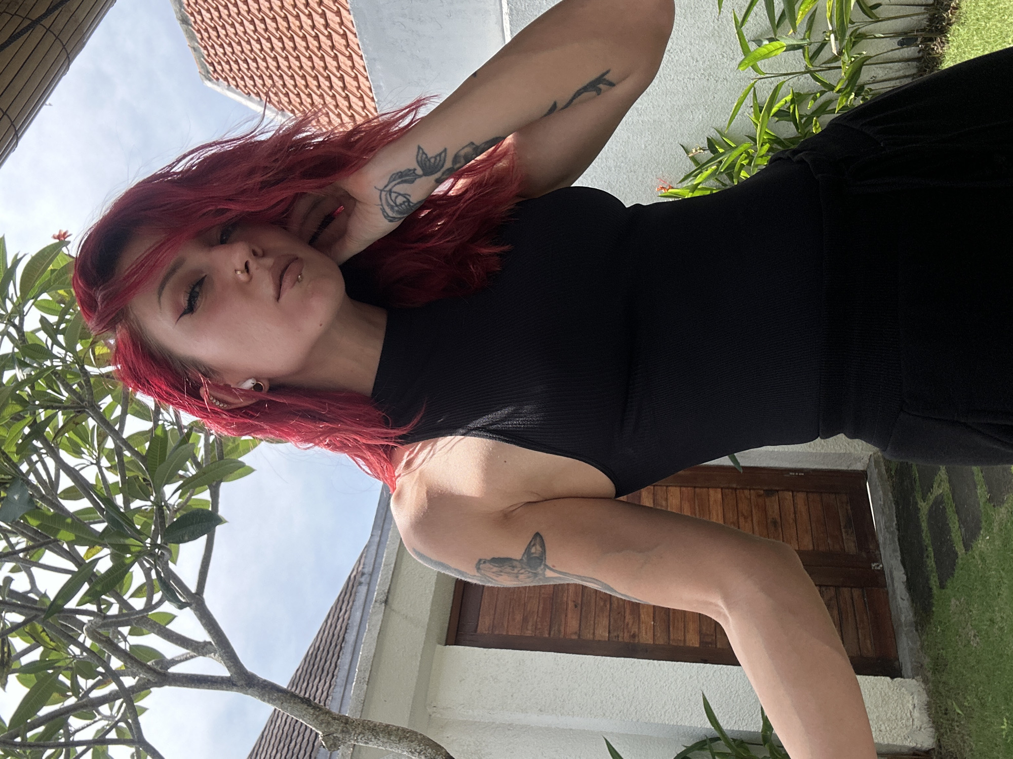 A woman with red hair, nose piercings, and a black outfit is depicted in a photograph. Her left arm is raised near her face, revealing a tattoo of a rose. A butterfly tattoo is visible on her right arm. The background includes a building with brown wooden doors, a white wall, plants, green grass, trees, and a section of a tiled roof. The setting is North Kuta, Indonesia.