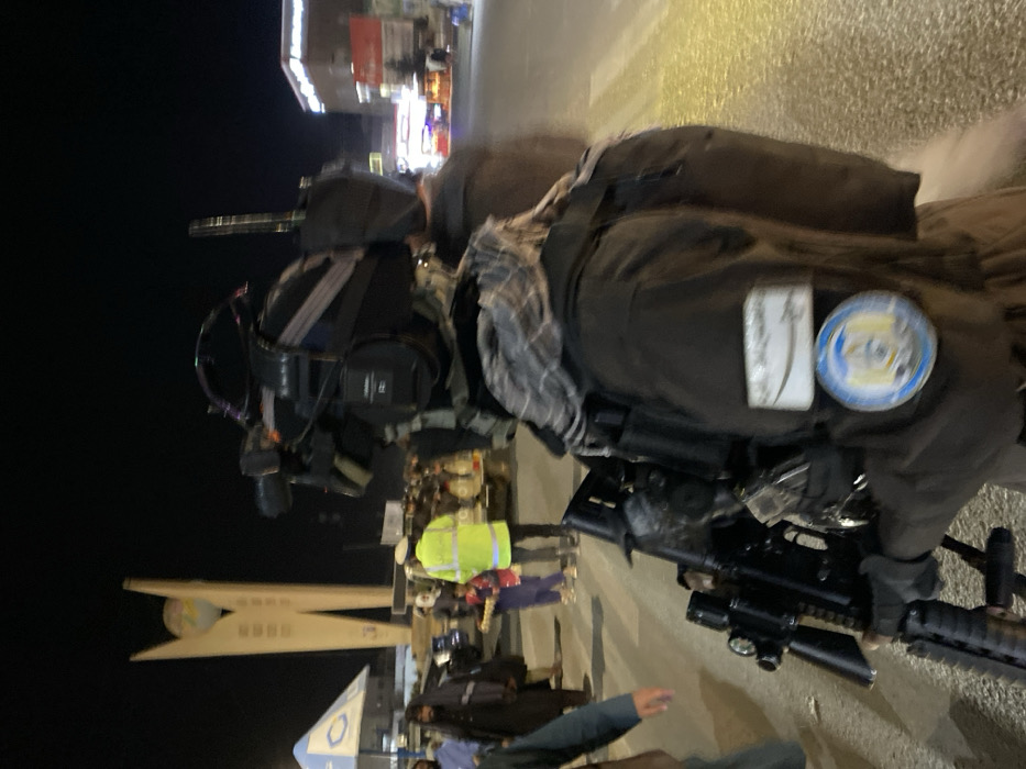 The image depicts a nighttime scene at Shohada Chowk (Martyrs' Roundabout) in Kandahar City, Afghanistan, centered around the distinctive Martyrs' Monument with its globe structure. In the foreground, a person viewed from the back-right wears dark tactical clothing, including a jacket, a patterned head covering (keffiyeh/shemagh), and carries an assault rifle equipped with an optical sight, a tactical light, and a vertical foregrip. A professional video camera system, possibly body-worn or helmet-mounted, is visible on their back/shoulder, along with communication gear and an antenna. Two patches are affixed to the right sleeve/vest: a rectangular one with Arabic script and a sword motif, and a circular one with an emblem.
The midground features numerous individuals, including a person in a bright yellow high-visibility vest and cap, possibly directing traffic or providing security. A child in blue clothing holds a tray of items. Other civilians, primarily men in traditional Afghan attire, are present on the street and sidewalk. Vehicles, including a white truck/SUV, are visible. The street has clear crosswalk markings. Buildings with illuminated signs line the background. The area is illuminated by artificial streetlights, indicating an active urban environment after dark.