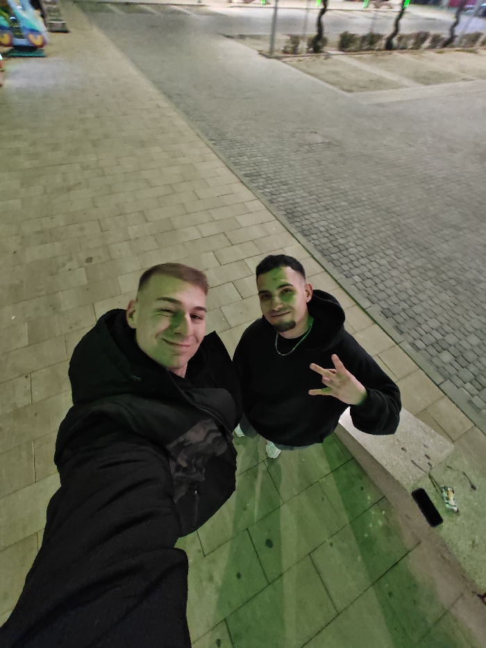 The image depicts two males posing for a selfie at night in an urban outdoor paved area in Shumen, Bulgaria.
The individual on the left, closer to the camera, is a male with light skin, short light-colored hair, and a clean-shaven face. He wears a dark hooded jacket over a patterned shirt and smiles while winking his left eye. His right arm is extended, holding the camera for the selfie.
The individual on the right, positioned slightly behind the first, is a male with light skin, dark short hair, and a short beard. He wears a black hooded sweatshirt and a silver-colored chain necklace. He smiles faintly and makes a "rock-on" hand gesture with his right hand.
The foreground ground is paved with light-colored rectangular flagstones. To the right of the individuals, there is a low, light-colored stone wall or ledge. Resting on this ledge are a black rectangular smartphone, a small white object (possibly a lighter or packet), and a discarded white cigarette butt.
In the mid-ground, the paving transitions to darker, smaller paver stones or cobblestones. In the far background to the left, a portion of a colorful children's amusement ride (blue, yellow, pink) is visible. Further back and to the right, bare-branched trees are visible alongside faint artificial lights, suggesting a street or parking area beyond the paved pedestrian zone.
The scene is illuminated by artificial light, casting a distinct green hue on the individuals' faces and the ground in the immediate foreground, indicating a specific light source. Shadows are cast beneath the individuals, suggesting an overhead or front-facing light source.