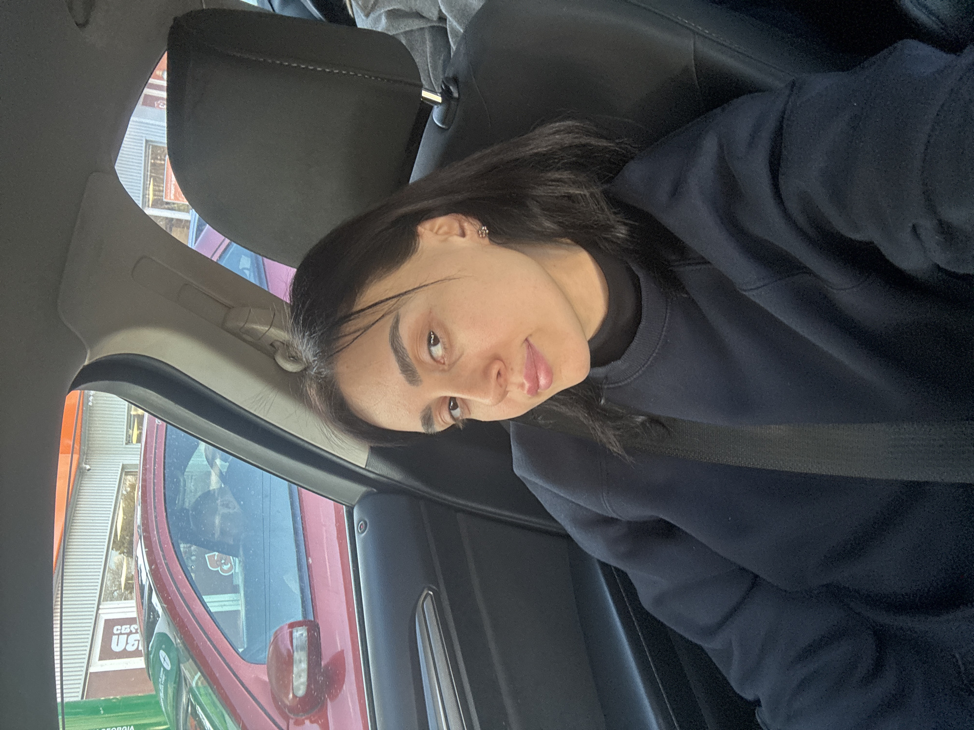 A woman wearing a navy sweater is sitting in the driver's seat of a car, wearing a seatbelt. Outside the car window, there is a building with a sign that says "CAS US GEORGIA" indicating the location is in Tbilisi, Georgia. A red car is parked next to the car the woman is in.