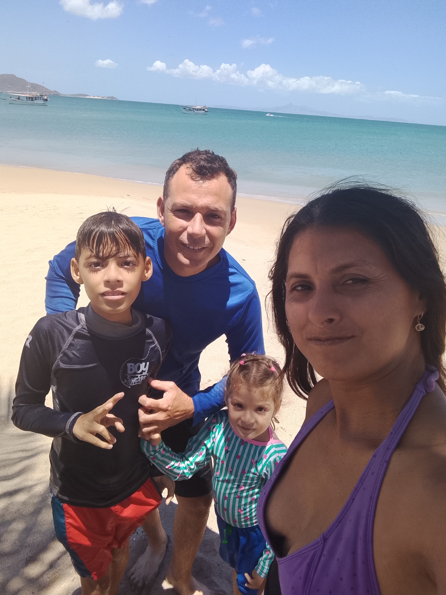 This vibrant, sunlit photograph captures a family of four posing on a beach in Juan Griego, Venezuela. The image features two adults and two children, positioned closely in the foreground, suggesting a group photo or selfie.
On the left, a young boy in a black long-sleeved rash guard with "BOY Surfer FISHING" text and red and blue swim shorts makes a hand gesture. Behind him, an adult man in a blue long-sleeved shirt smiles warmly at the camera. To the right, an adult woman in a purple halter-neck swimsuit top looks directly at the viewer with a subtle smile. Peeking from behind her, a small girl in a green and white striped long-sleeved top with pink stars and blue shorts also looks towards the camera; faint text resembling "HAWAI" is visible on her top.
The setting is a picturesque beach with light-colored sand in the foreground, leading to clear, turquoise-blue ocean water under a bright blue sky with scattered white clouds. In the mid-ground, two small boats are visible on the calm water, with distant hills or mountains defining the coastline. The clear skies and bright illumination indicate a sunny day, likely midday. The overall atmosphere is relaxed and joyful, capturing a pleasant family moment by the sea.