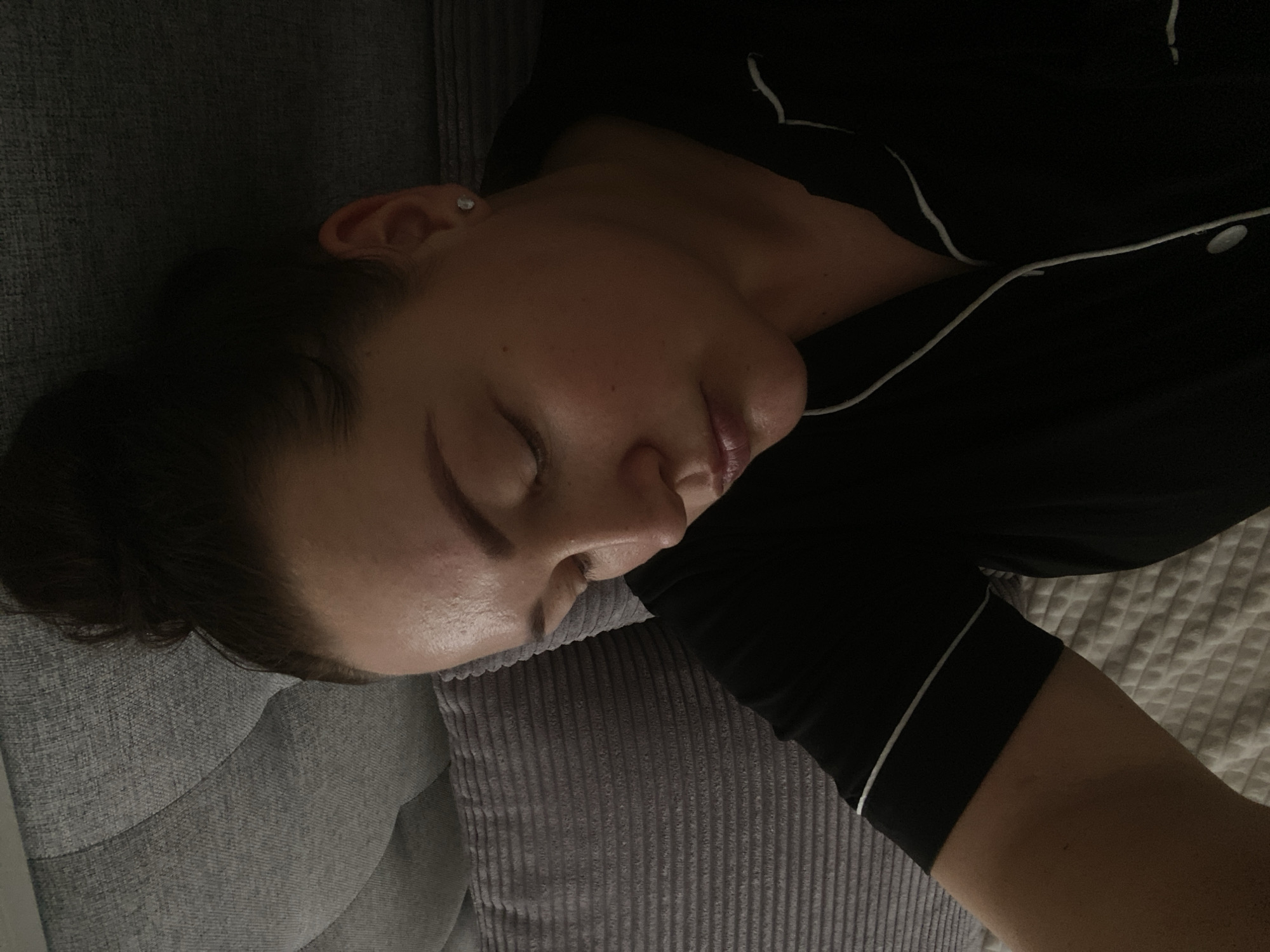 A woman is asleep on a grey couch in what appears to be a residential setting. She is wearing a black top with white piping, possibly pajamas. Her hair is pulled back in a bun. The background shows a bed with a white comforter. There is no indication of the location being Sochi, Russia.