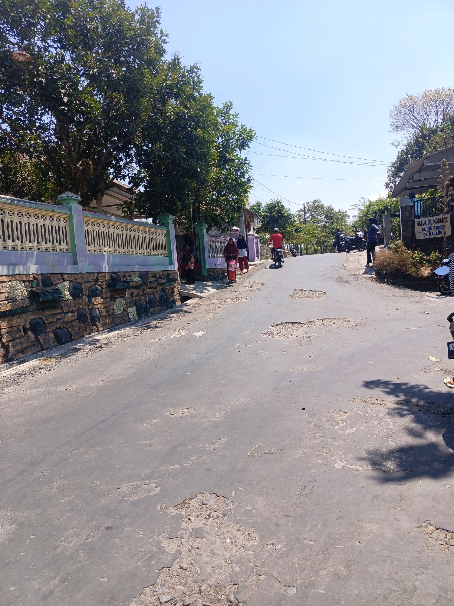 The image depicts a road in Kalang, Indonesia, characterized by its poor condition with numerous potholes and cracked asphalt. The road runs uphill and curves to the right. On the left side of the road, a decorative stone wall with a patterned fence runs along the property line. Trees with dense foliage are visible behind the fence. On the right side of the road, a sign reads "MASUK DK. NGRAMBU RT.008 RW.009 DS. KALANG", indicating the entrance to Ngrambu Hamlet in Kalang Village. Several people are present: two women in hijabs and one person in a red shirt on a motorbike are walking or riding on the road near the fence. Further up the road, a person in a blue shirt is standing, and several motorbikes are parked or being ridden. The sky is clear and blue, suggesting a sunny day. The overall scene portrays a rural or semi-rural road in Indonesia.