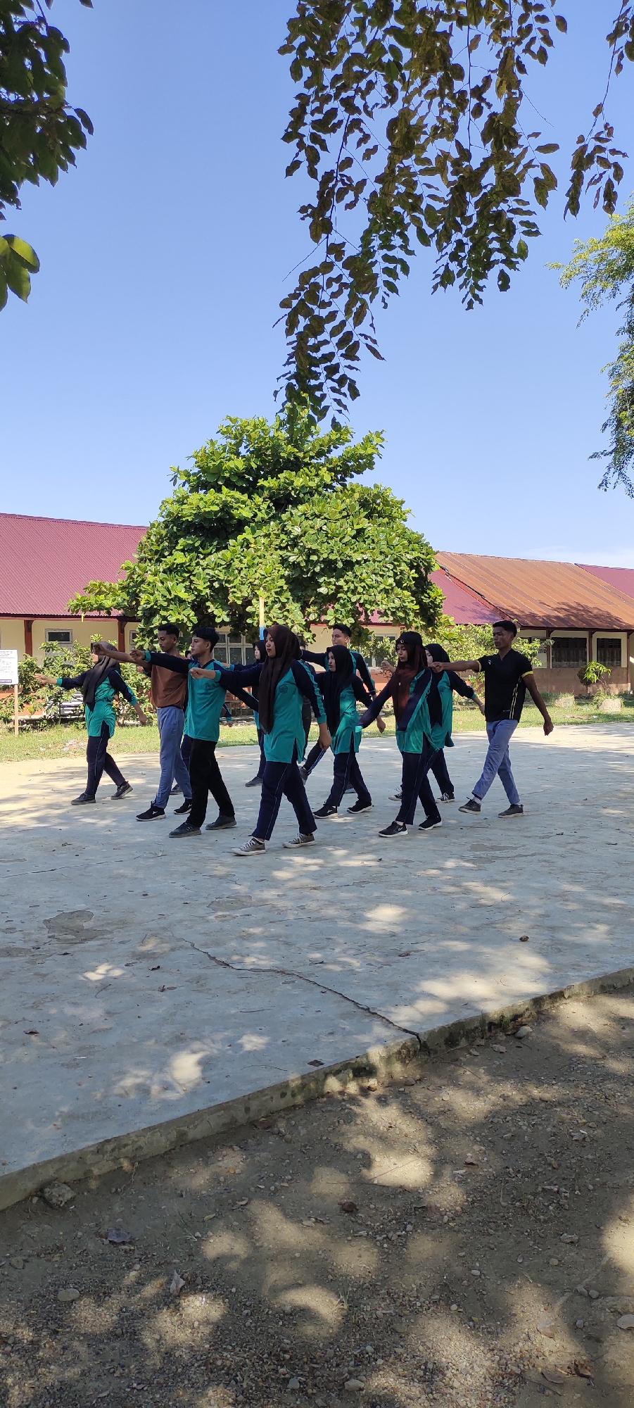 A group of nine individuals, consisting of six females and three males, are engaged in what appears to be a synchronized outdoor activity or exercise. They are positioned in a line, facing left and executing similar arm movements. The females are predominantly wearing teal shirts with dark blue accents and dark blue pants, with several also wearing dark headscarves. The males are dressed in a mix of teal shirts, dark blue pants, and one individual is in a brown shirt with dark pants.
The setting is an open, paved area with visible cracks in the concrete. In the background, there are buildings with red roofs and light-colored walls, suggesting an institutional or school-like environment. A large, leafy tree dominates the middle ground, with branches and leaves extending towards the top of the frame. The sky is clear and blue, and the scene is illuminated by sunlight, casting shadows on the ground. The overall impression is that of an organized outdoor activity taking place on a sunny day. The specific location of Langnibong, Indonesia, is mentioned as the city where this scene is situated.