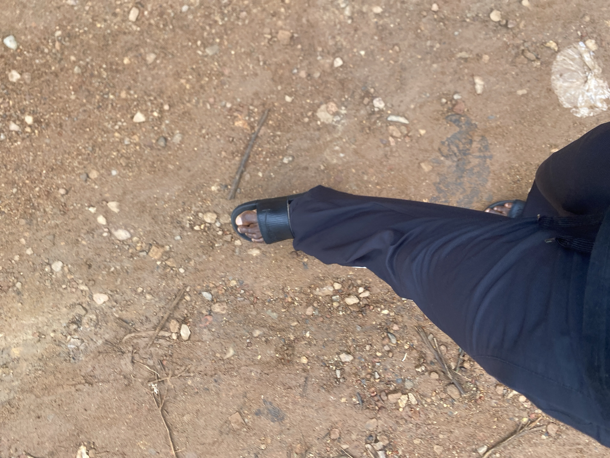 The image shows a first-person perspective of a person walking on a dirt path in Jos, Nigeria. The ground is covered in reddish-brown dirt, small stones, and scattered twigs. The person is wearing dark-colored pants and black slide sandals. The left foot and lower portion of the left leg are visible. A discarded plastic bag is visible on the right side of the path. The lighting suggests it is daytime.