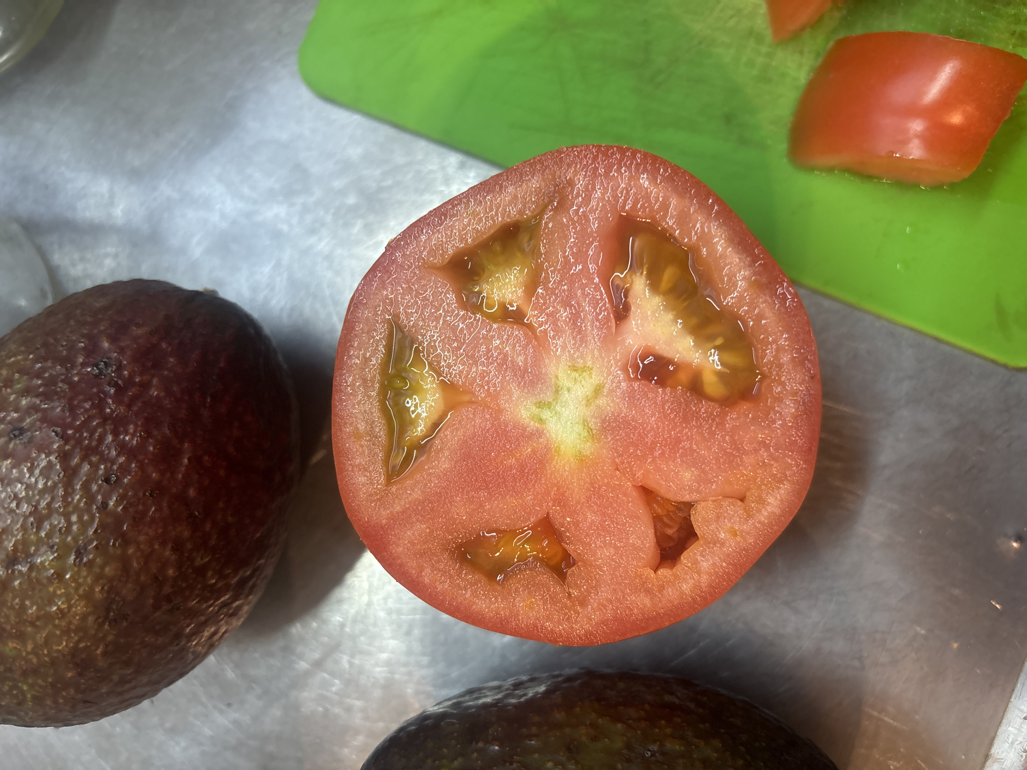 The image shows a close-up of food preparation. A cross-section of a tomato is prominently displayed, revealing its inner structure and seed cavities filled with liquid. Adjacent to the tomato is an avocado, with its dark, bumpy skin visible. Part of a green cutting board is also in the frame with sliced tomato sitting on it. The food items are placed on a surface, possibly a metal countertop. The composition suggests a meal preparation scene. There is no information that indicates the location of the picture to be in Buenos Aires, Argentina.