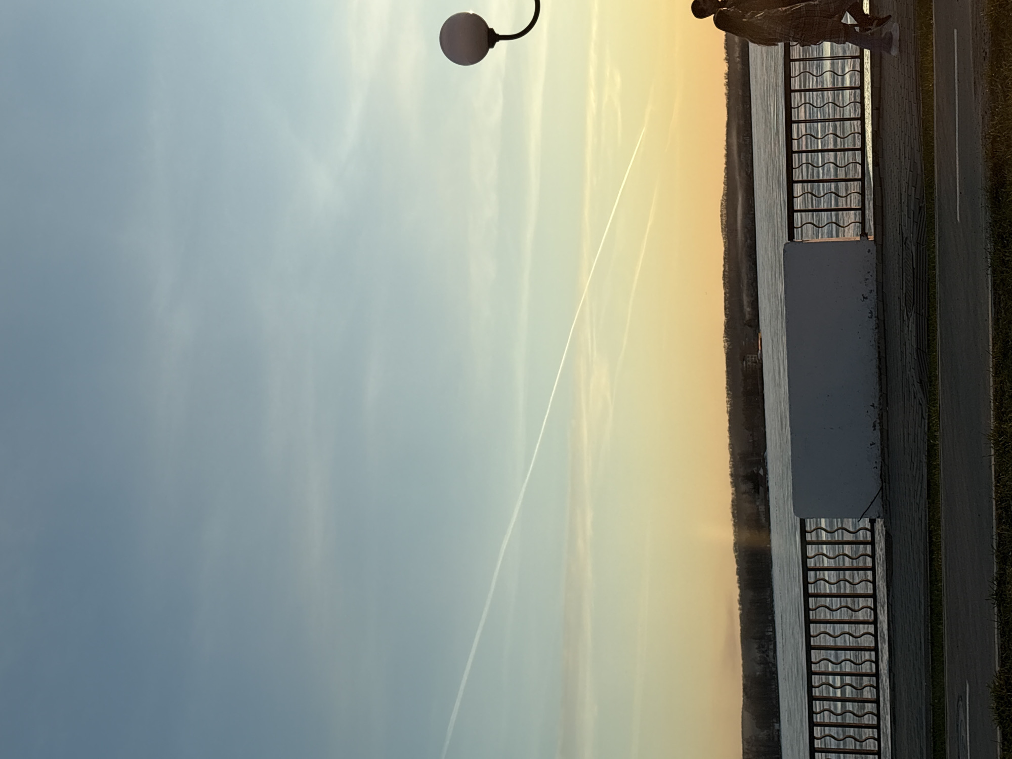 This image captures a serene waterside scene, likely along a river or large lake in Votkinsk, Russia, at either sunrise or sunset. The sky displays a beautiful gradient, transitioning from a soft, clear blue on the left to a warm, golden yellow and orange on the right, indicative of the golden hour. A distinct, straight white contrail from an aircraft cuts diagonally across the sky.
In the foreground, a paved promenade or pathway runs alongside the water. A black metal railing with a decorative wavy pattern, mounted on a white concrete barrier, separates the path from the expansive body of water. A single person is visible on the promenade, standing near the railing and looking out over the water towards the distant, dark shoreline, which appears to be lined with trees or distant landmass. The individual is dressed in a plaid-patterned top and dark pants. A modern street lamp with a spherical white light fixture is partially visible in the upper left corner of the frame. The water itself reflects the soft light of the sky. No text is visible in the image.