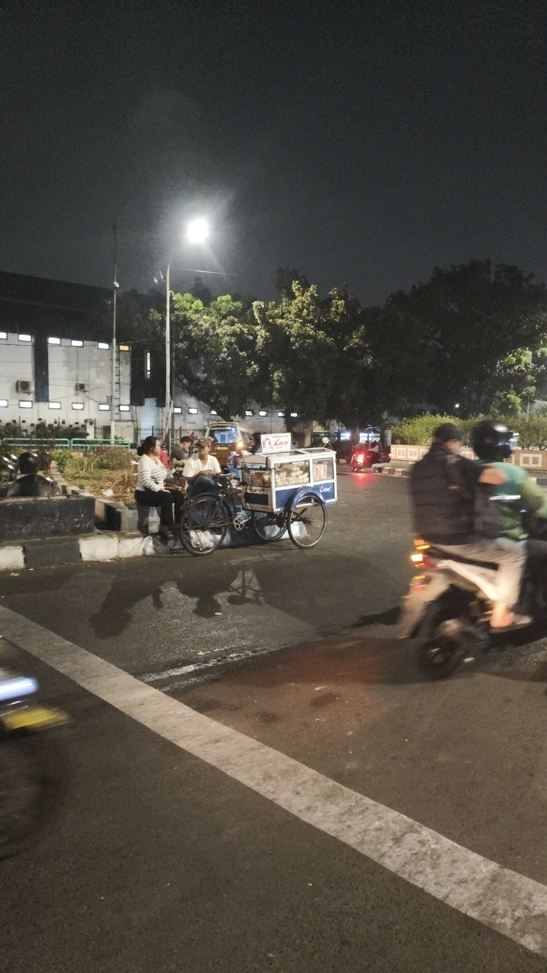The image depicts an outdoor scene at night, likely in a public area of South Jakarta, Indonesia. A street lamp illuminates the area. In the foreground, a wet asphalt road with a white line marking is visible, with a blurred yellow object on the left side.
In the mid-ground, a food cart on wheels, possibly selling snacks or drinks, is parked on the side of the road. Two individuals are seated on a concrete barrier next to the cart. One person, a woman wearing a striped long-sleeved shirt and dark pants, is seated and looking towards the cart. The other person, a man wearing a white t-shirt and dark pants, is seated and appears to be interacting with the cart.
To the right of the food cart, two people are riding a motorcycle. The person in the front is wearing a dark jacket and a helmet, while the person in the back is wearing a green jacket and a helmet. They are moving away from the camera.
In the background, there is a building with multiple windows and lights, suggesting it might be a commercial or institutional building. Trees with dense foliage are also visible, and further in the distance, there are other lights from vehicles and potentially more buildings, indicating an urban environment. The overall atmosphere is that of a busy street at night with some commercial activity.