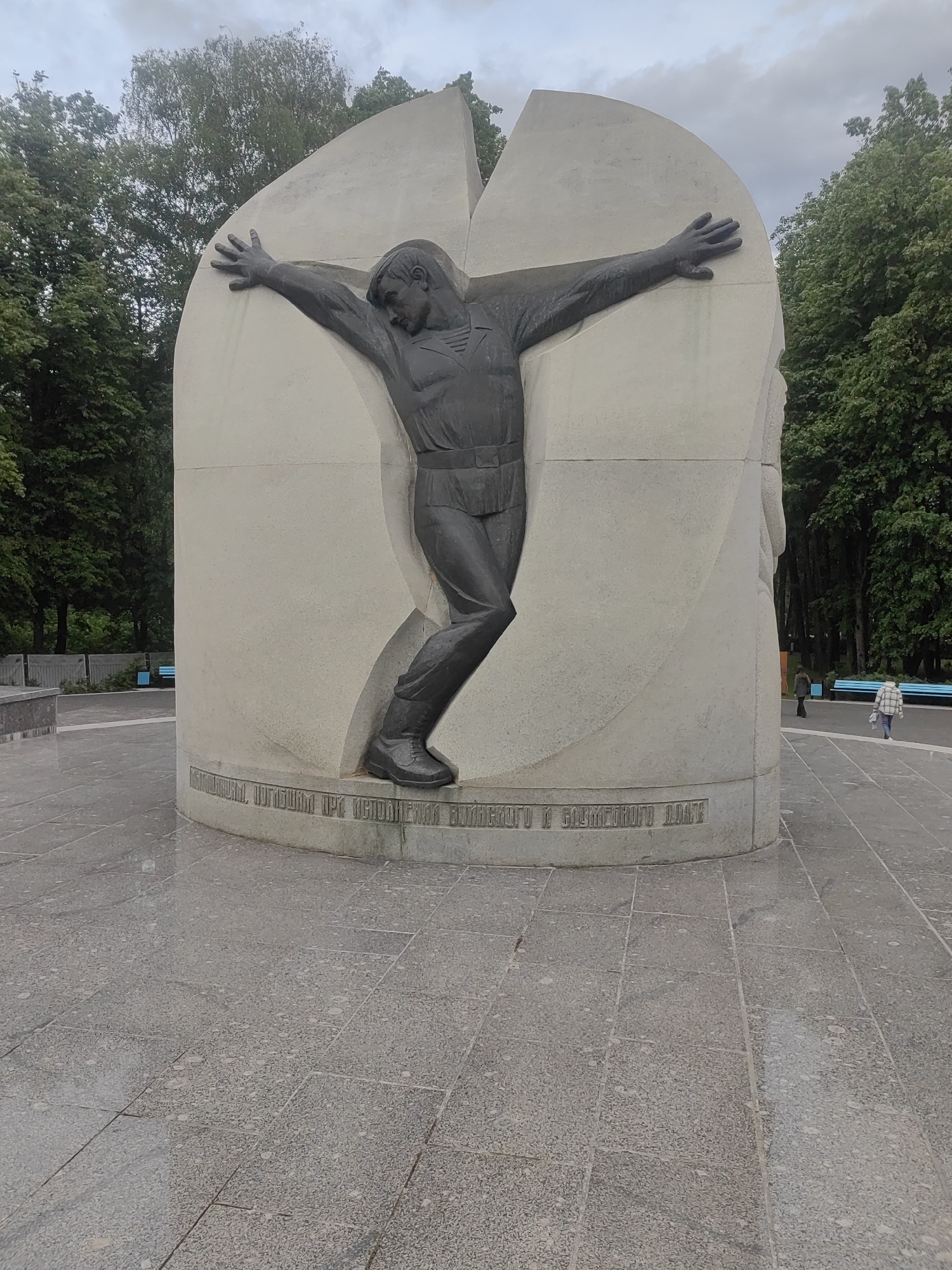 The image shows a monument dedicated to the victims of the Chernobyl disaster in Mytishchi, Russia. The monument features a dark metal sculpture of a man with outstretched arms, embedded in a light-colored stone structure that appears to be cracked open. The man is depicted in what seems to be work clothes, including a shirt and trousers, with work boots on. The monument is placed on a circular base with an inscription in Cyrillic. The surrounding area is paved with gray stone tiles. In the background, there are green trees, suggesting a park setting. There are benches in the background and people are walking in the park. The sky is overcast.