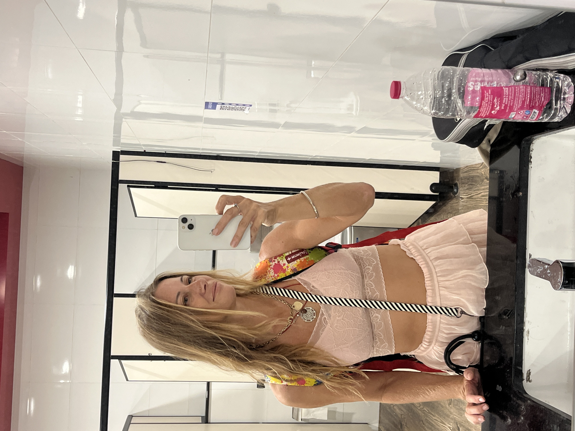 This media file shows a woman taking a mirror selfie in what appears to be a public restroom. She has long blonde hair and is wearing a pink lace bralette and a light pink ruffled skirt. She is also wearing a necklace with multiple pendants and a colorful lanyard across her chest. In the reflection of the mirror, a plastic bottle of water with a pink label is visible, along with a black bag. The walls of the restroom are tiled with white, glossy tiles. The overall impression is that of a casual moment captured by the subject. The location is identified as Katunayake, Sri Lanka.