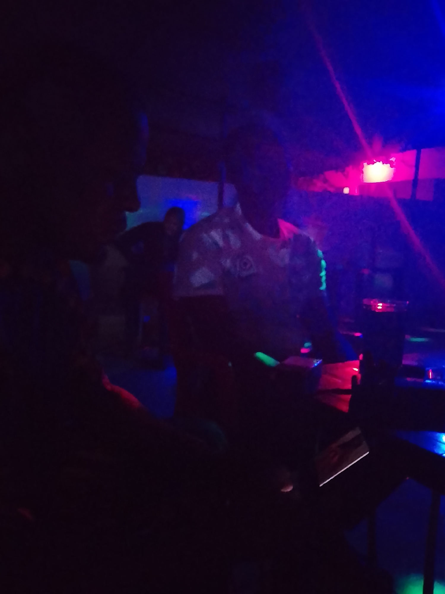 The image captures a dark, indoor scene bathed in vibrant artificial light, likely a nightclub, bar, or party venue. The primary light sources are bright blue, pink/red, and some green neon or UV lights, creating a high-contrast, energetic atmosphere.
In the foreground, a person is visible on the left, largely in silhouette, facing right. Another person stands in the center, wearing a light-colored short-sleeved shirt that reflects the ambient lights. This person appears to be interacting with a setup of equipment, strongly resembling a DJ mixer or sound board, which is adorned with numerous glowing red, blue, and green lights. Notably, there are also some distinct neon green glowing elements on the central person's right arm, possibly a bracelet or design on the sleeve. A third, more distant figure can be faintly seen in the background, further to the right.
The activity taking place appears to be a social gathering or event where music is being played, with the central figure possibly acting as a DJ or technician. The time of day is undoubtedly nighttime, given the reliance on artificial illumination. No specific weather conditions are discernible from this indoor shot. There are no clear location cues beyond the nightclub/party setting. The overall mood is lively and atmospheric. No discernible text is visible in the image.