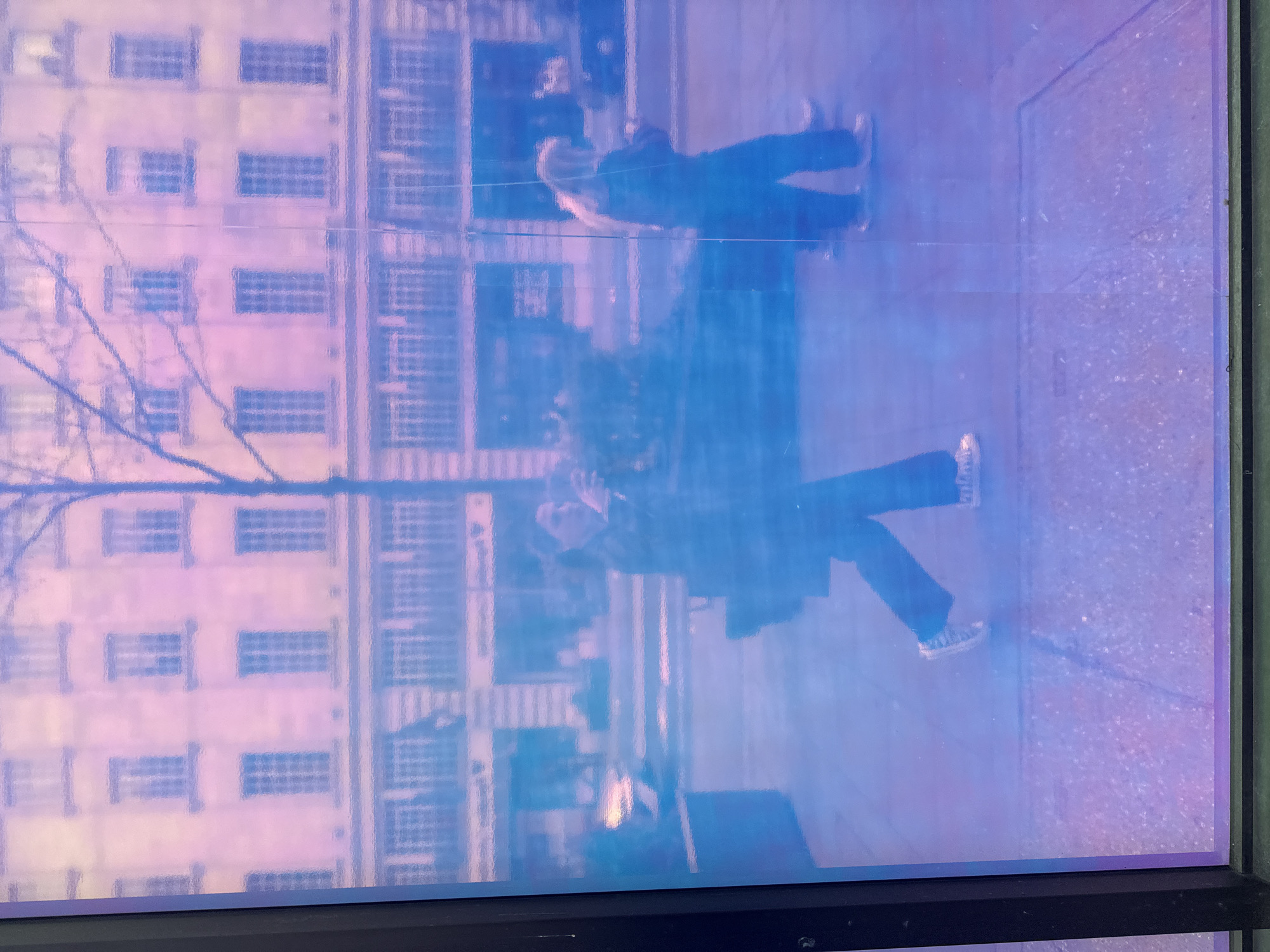 The image shows a reflection in a window. The reflection shows a building with many windows and what appears to be a tree. Two people are also reflected in the window. The window is likely located in London, United Kingdom.