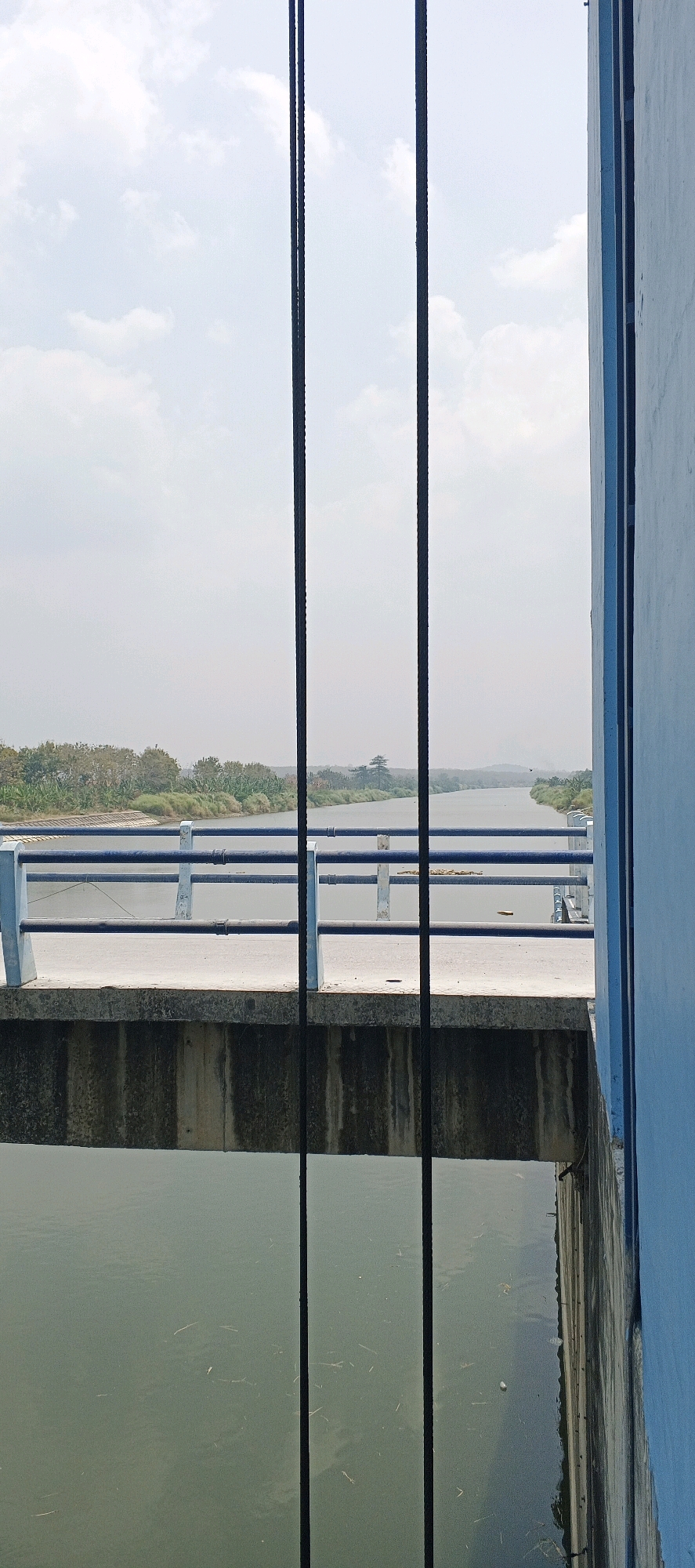 The image captures a view from a concrete structure overlooking a calm river. The foreground is dominated by the dull grey concrete of the structure, with darker, vertical lines suggesting cables or guide rails. Below this structure, the murky green water of the river is visible, with scattered debris. In the mid-ground, a bridge or walkway extends across the river, featuring a blue railing with white support posts. Beyond the bridge, the river curves to the left, bordered by lush green vegetation and trees. The background consists of a hazy sky with scattered white clouds. The scene is devoid of any people or discernible activity, suggesting a still, natural setting within the industrial context of a river control or bridge structure. The location is identified as Parengan, Indonesia.